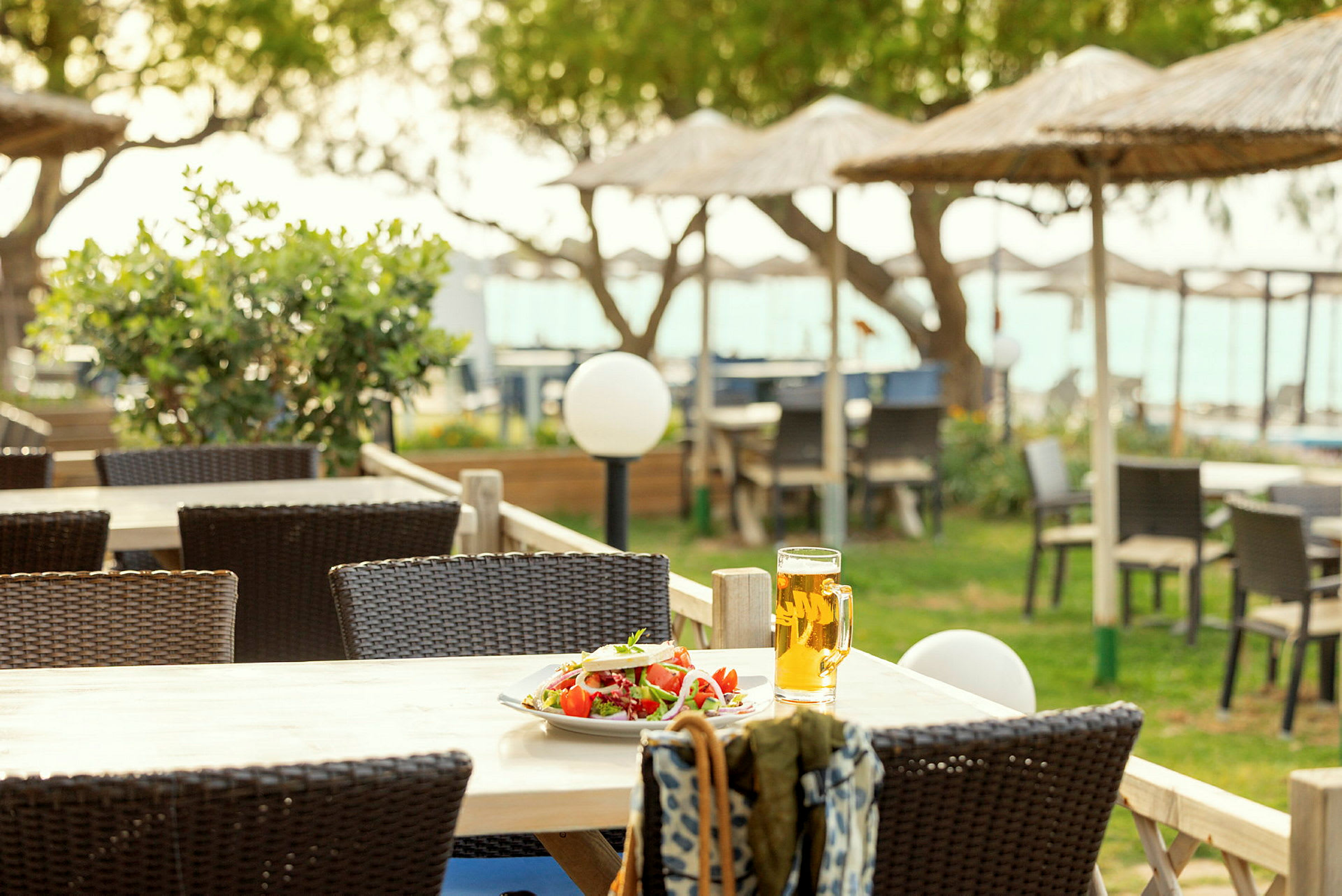 Et bord med en tallerken salat og et glass øl på en terrasse med utemøbler og strandparasoller i bakgrunnen. (AI-generert tekst)
