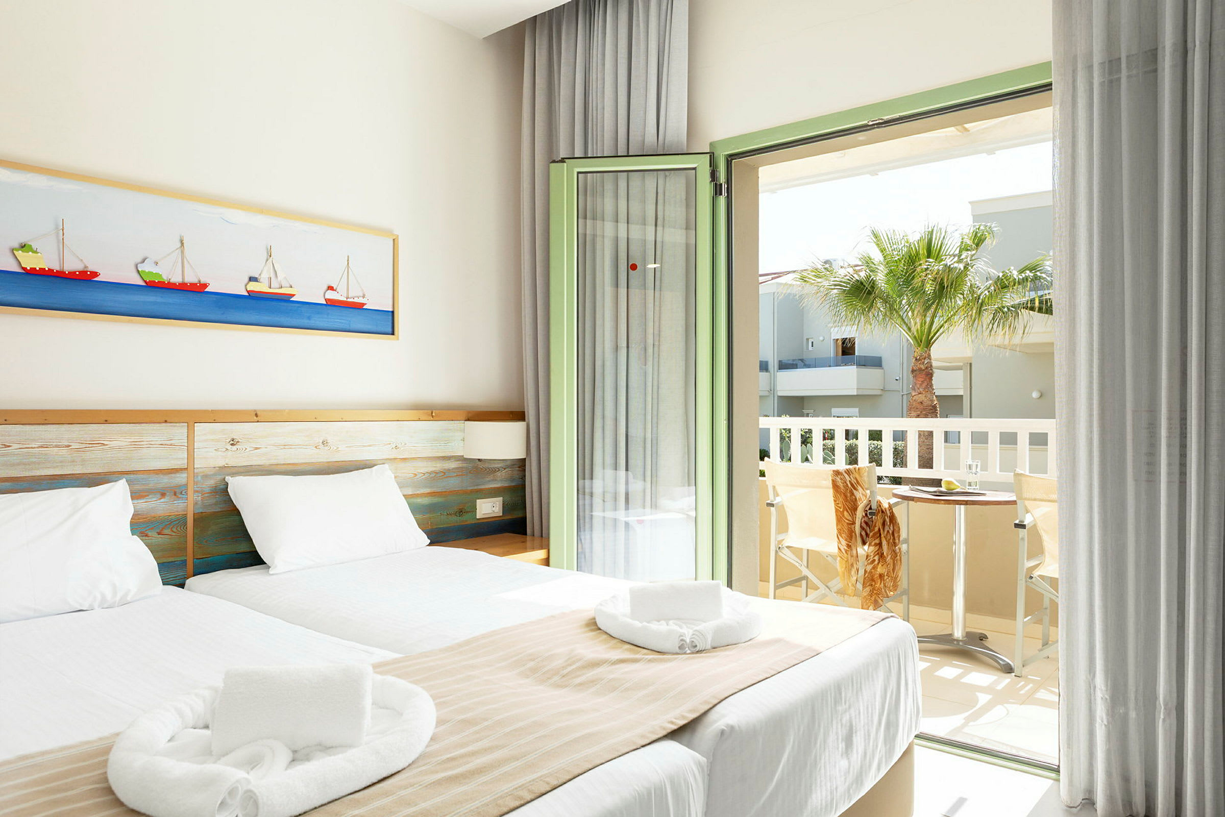 Et lyst hotellrom med to enkeltsenger, hvitt sengetøy og beige sengetepper. En kunst med båt motiver henger på veggen over sengen. Rommet har en åpen balkongdør med utsikt over en palme og et bord med to stoler. (AI-generert tekst)