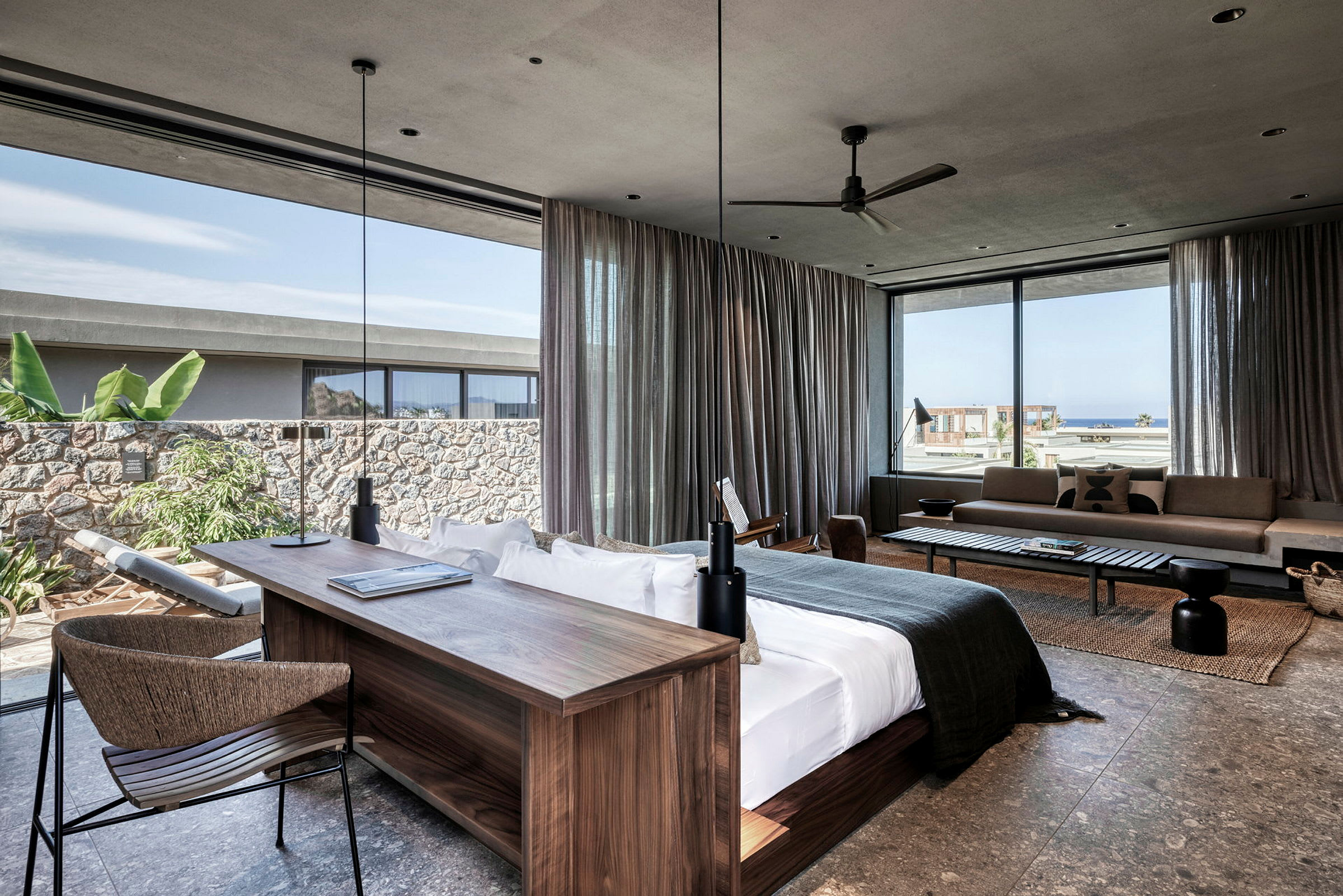 Moderne hotellsuite med stor seng, skrivebord og sitteområde. Vinduer med utsikt mot havet og vegetasjon. (AI-generert tekst)