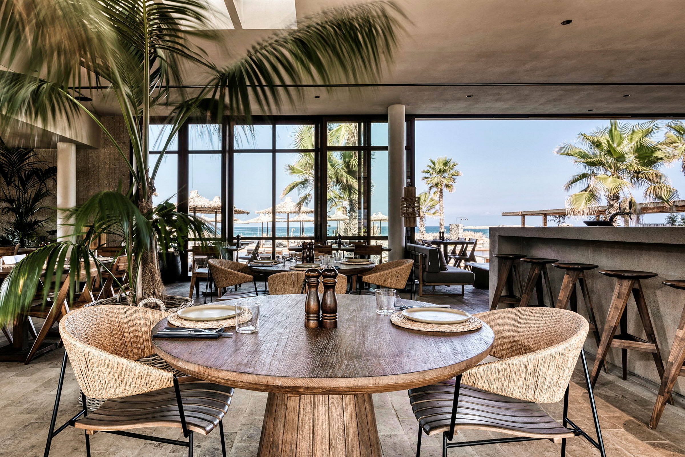 Restaurant med trebord og stoler, omgitt av palmer og havet i bakgrunnen. (AI-generert tekst)