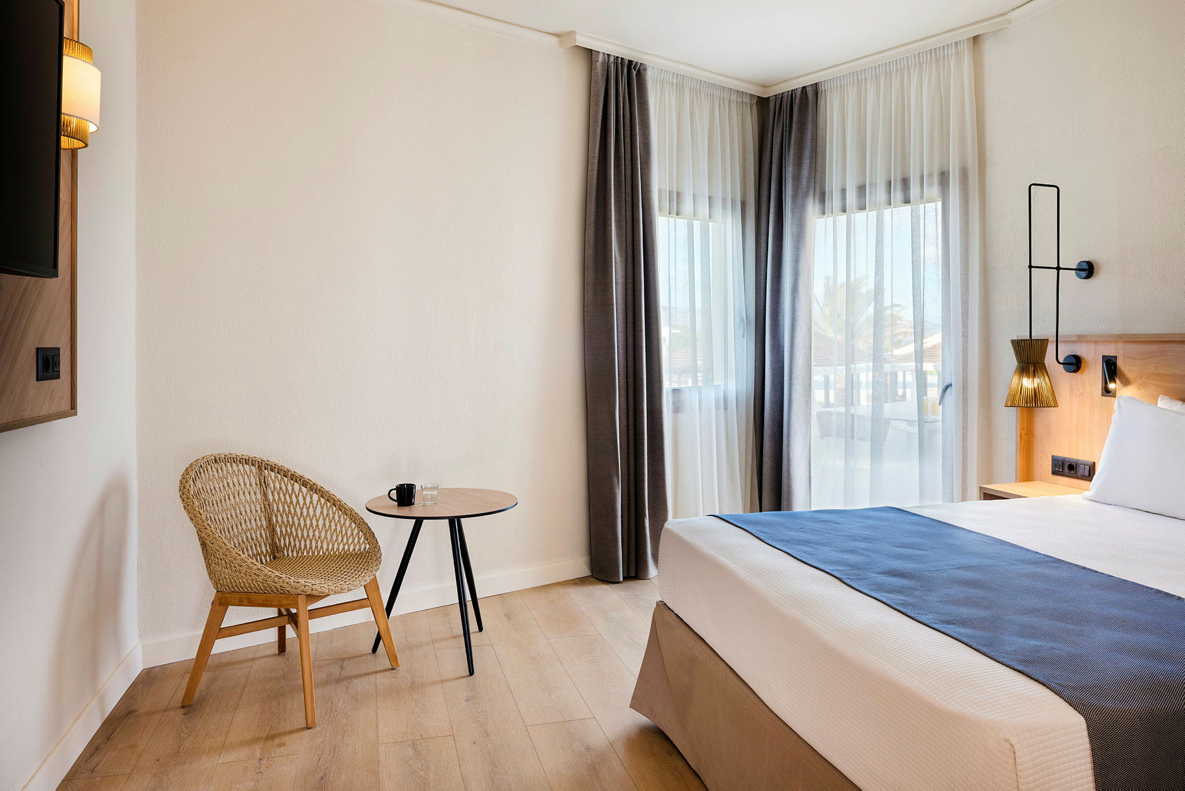 Et hotellrom med en moderne stol, et lite bord og en seng ved vinduer med lyse gardiner. (AI-generert tekst)