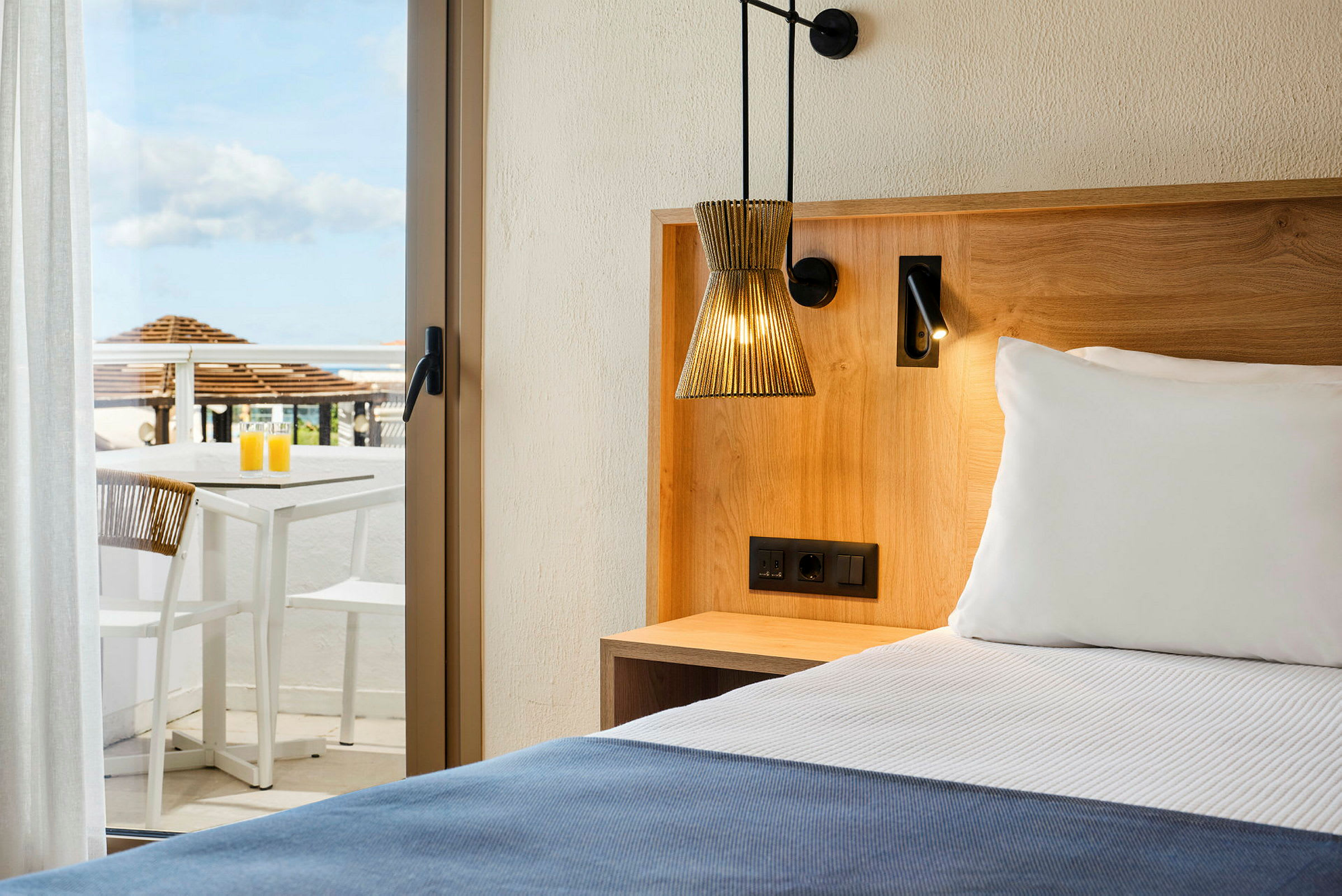 Hotellrom med en seng, treinnredning og en terrasse med bord og stoler i bakgrunnen. (AI-generert tekst)
