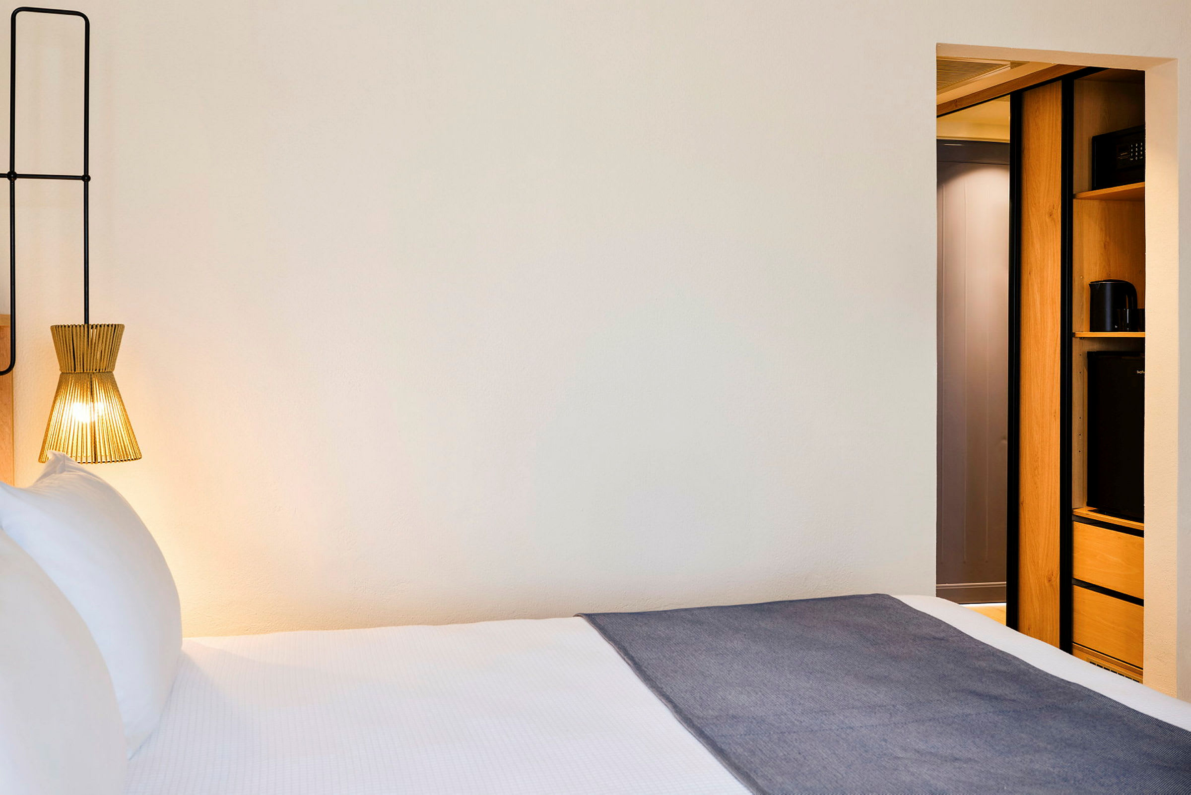 Et hotellrom med en seng, en moderne lampe og en del av innredningen med lagringsplass. (AI-generert tekst)