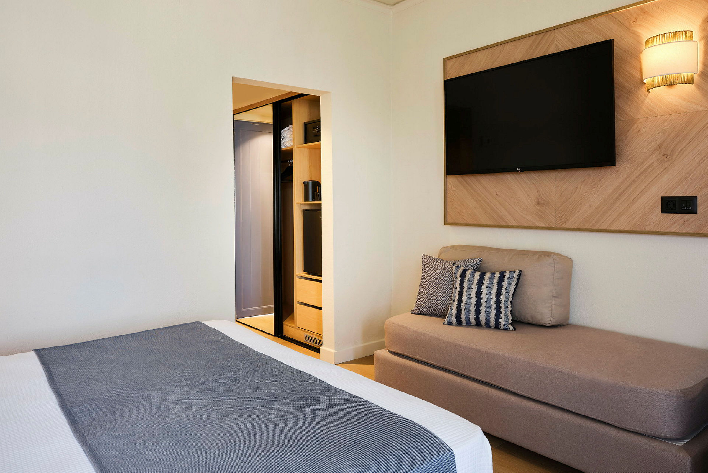 Et hotellrom med en seng, en sofa, en veggmontert TV og et åpent garderobeskap. (AI-generert tekst)