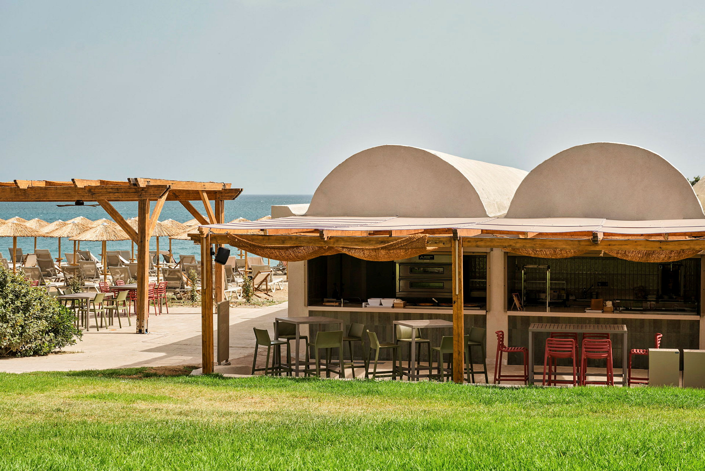 Utendørsrestaurant ved stranden med grønn gressplen, trepergola og parasoller. (AI-generert tekst)