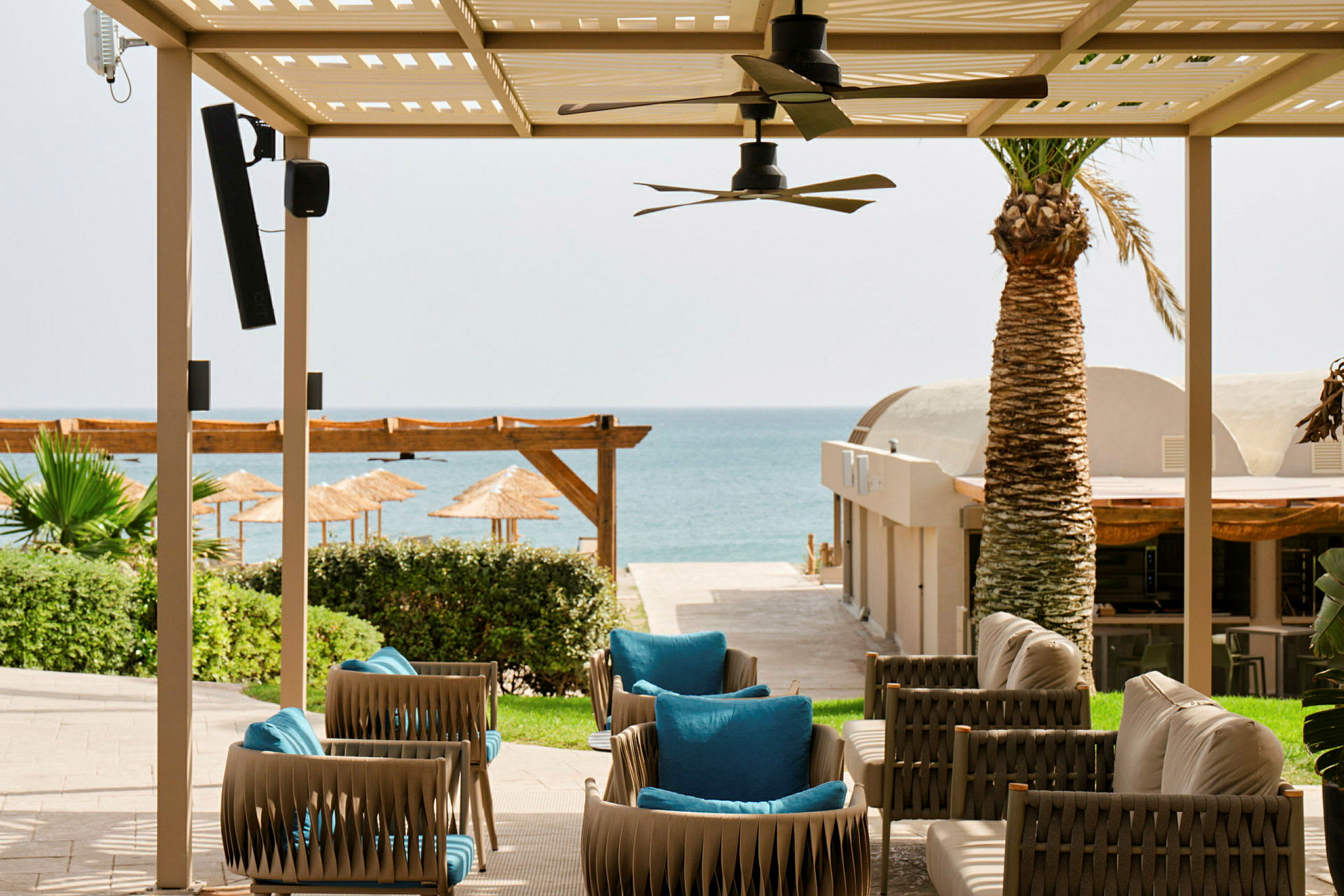 Hagemøbler under en pergola med utsikt mot en sandstrand og havet i bakgrunnen. (AI-generert tekst)