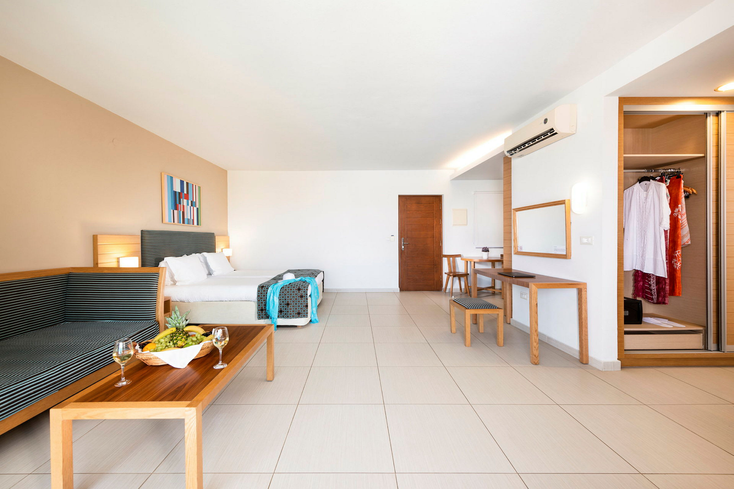 Et romslig hotellrom med dobbeltseng, sittegruppe, spisebord, speil og garderobe. En fruktskål og vinglass står på sofabordet. (AI-generert tekst)