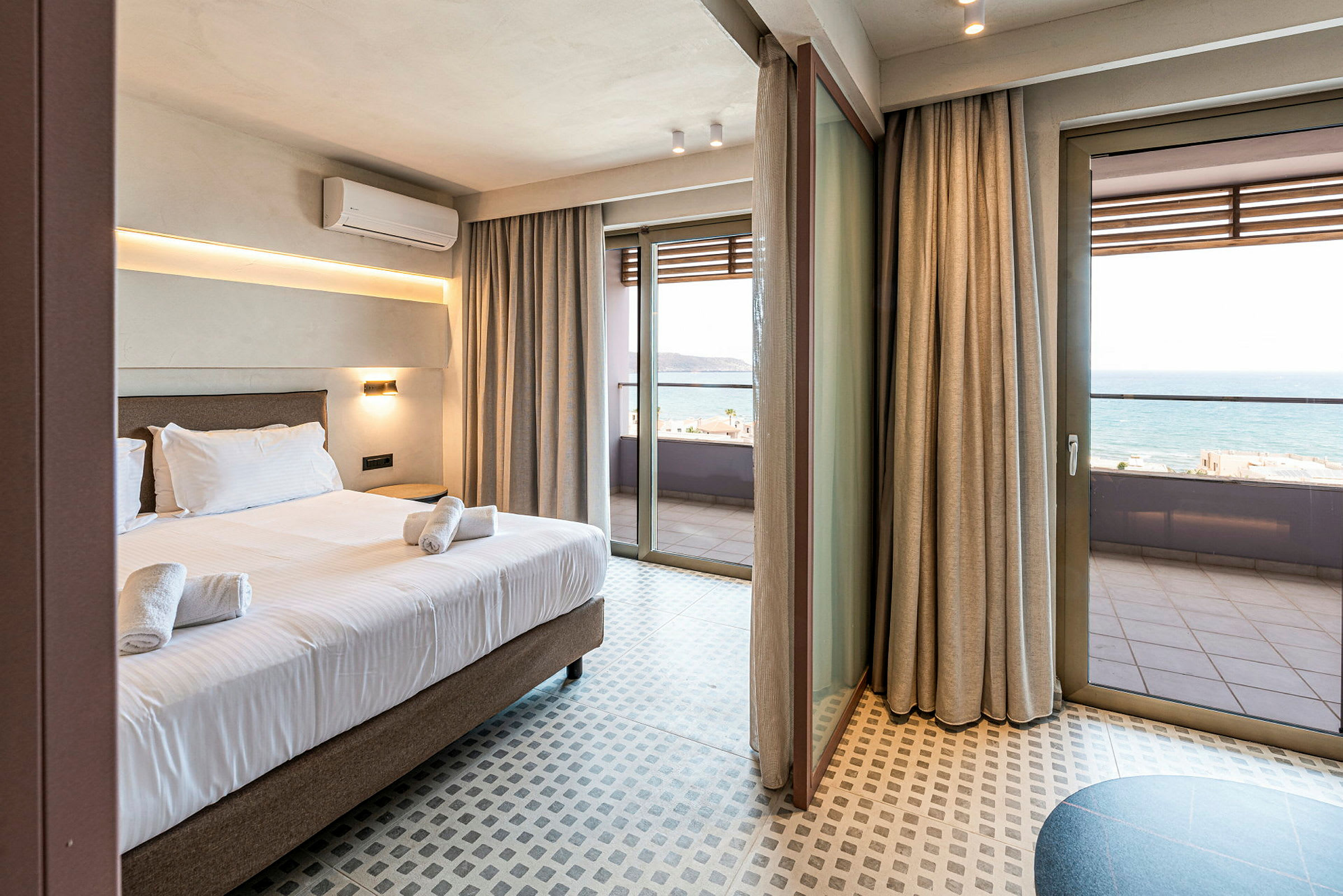 Hotellrom med seng og håndklær, store glassdører som fører ut til en balkong med havutsikt. (AI-generert tekst)