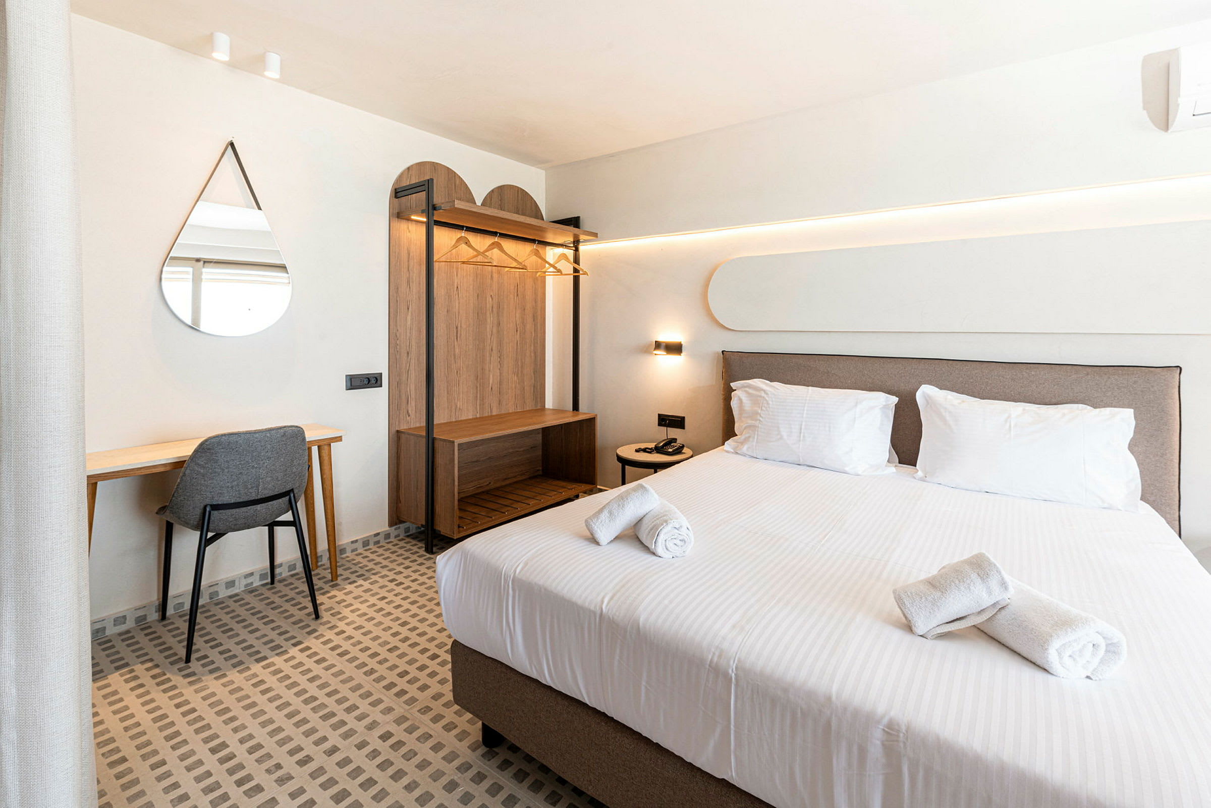 Et hotellrom med en oppredd seng, skrivebord med stol, veggspeil og åpen garderobe. (AI-generert tekst)
