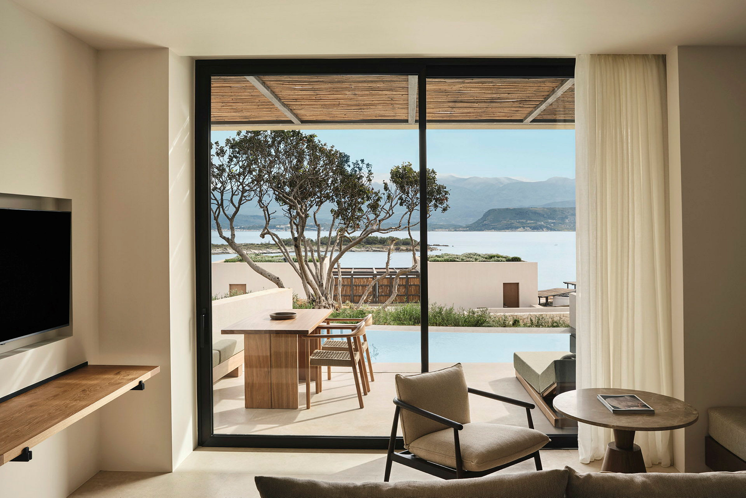 Utsikt fra et rom med moderne innredning mot en terrasse med bord og stoler, samt utsikt over havet og fjellene. (AI-generert tekst)