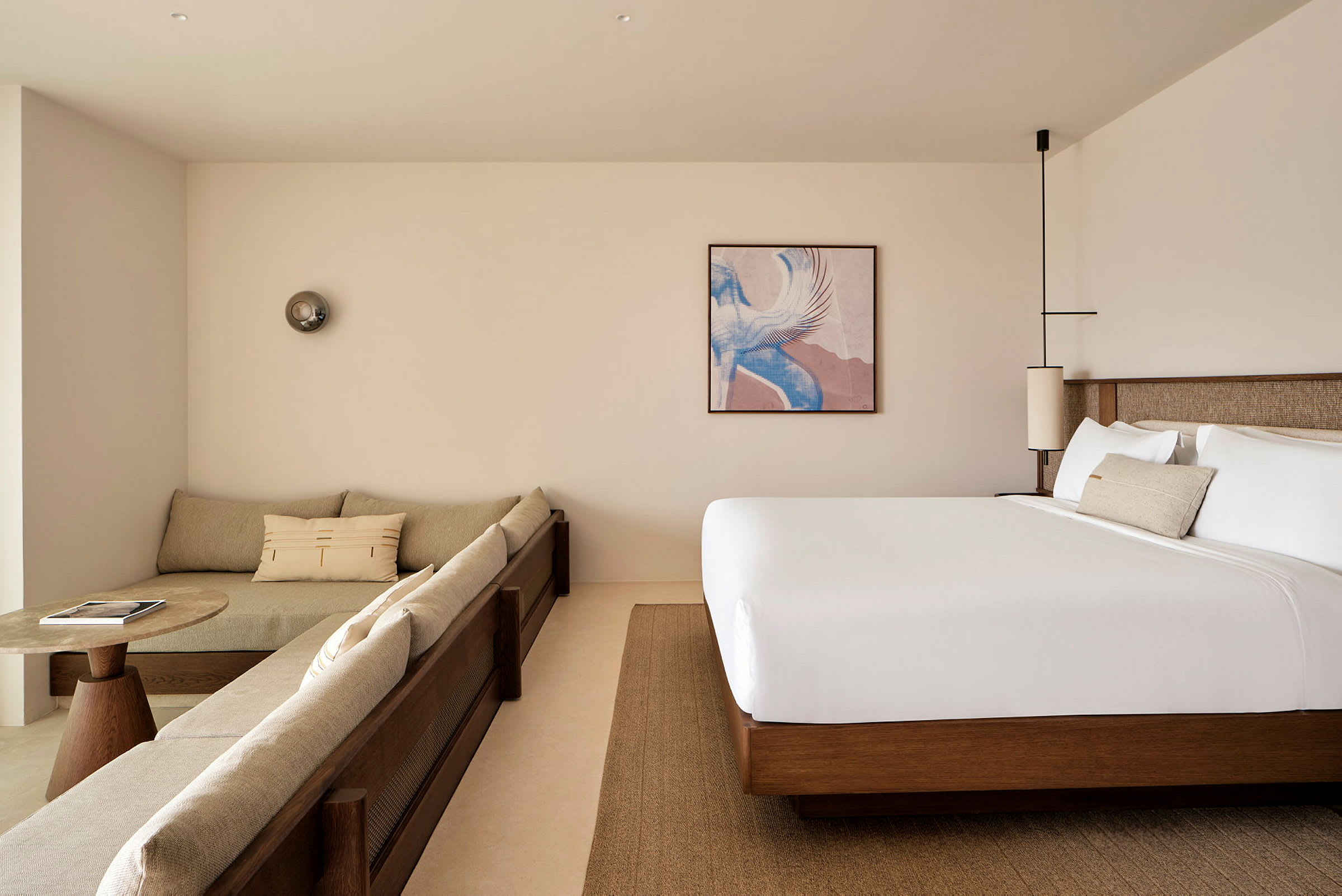 Et moderne hotellrom med beige sofa, et rundt sofabord og en stor seng. Et bilde samt dekorative lamper er synlige. (AI-generert tekst)