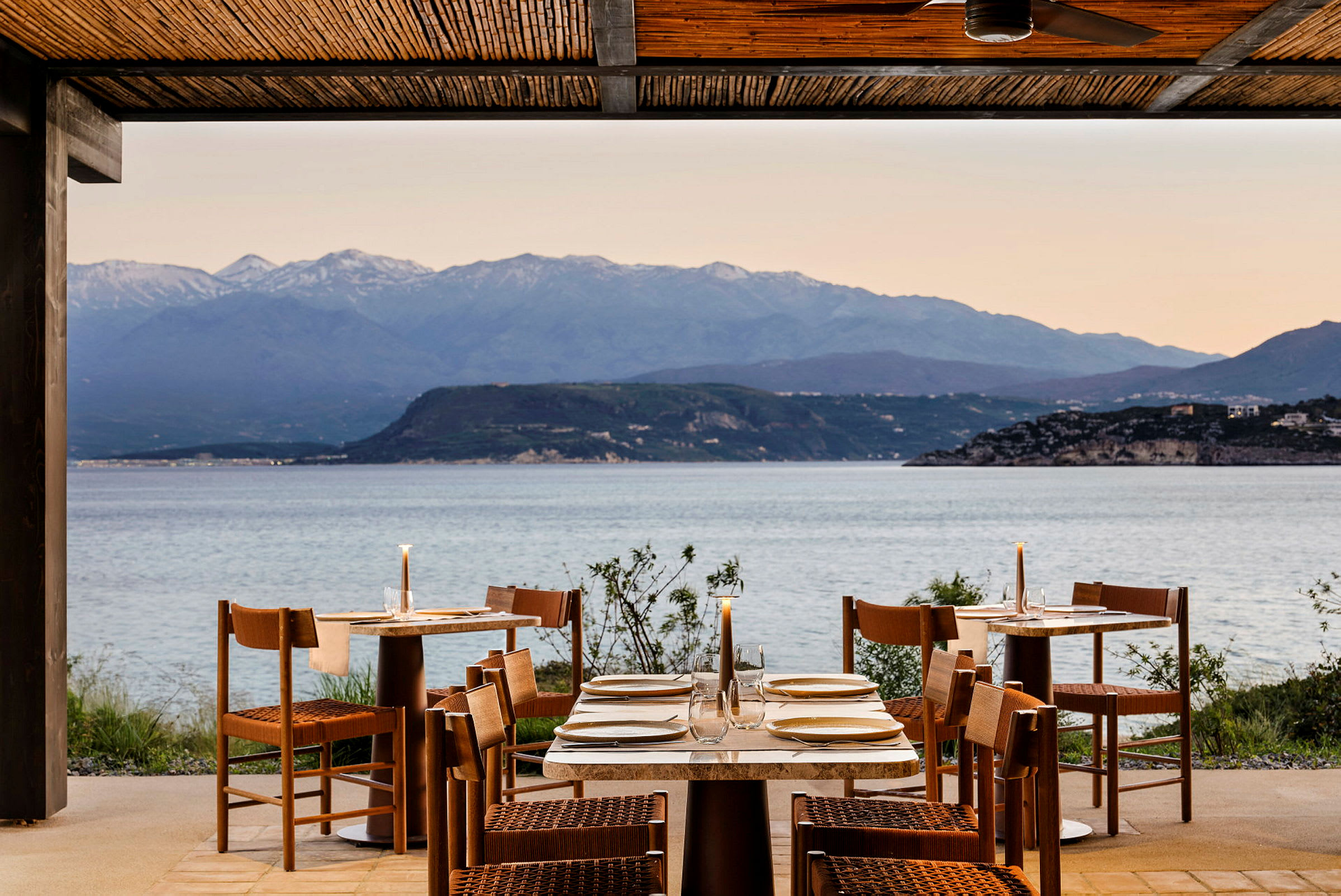 En utendørsrestaurant med dekkede bord, med utsikt over havet og fjell i bakgrunnen. (AI-generert tekst)
