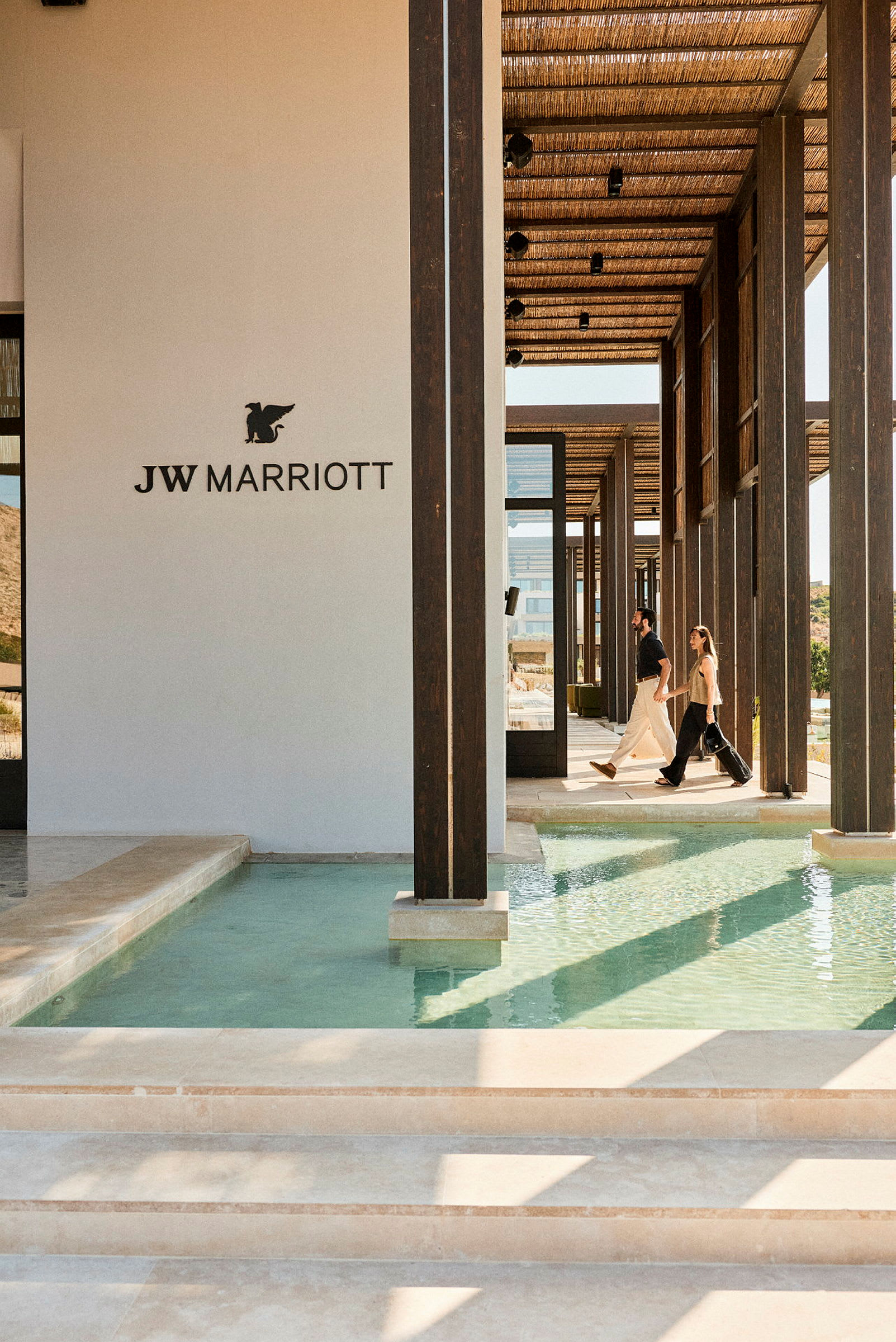 Bilde av en moderne bygning med JW Marriott-logoen, hvor folk går langs en gang med vann på begge sider. (AI-generert tekst)