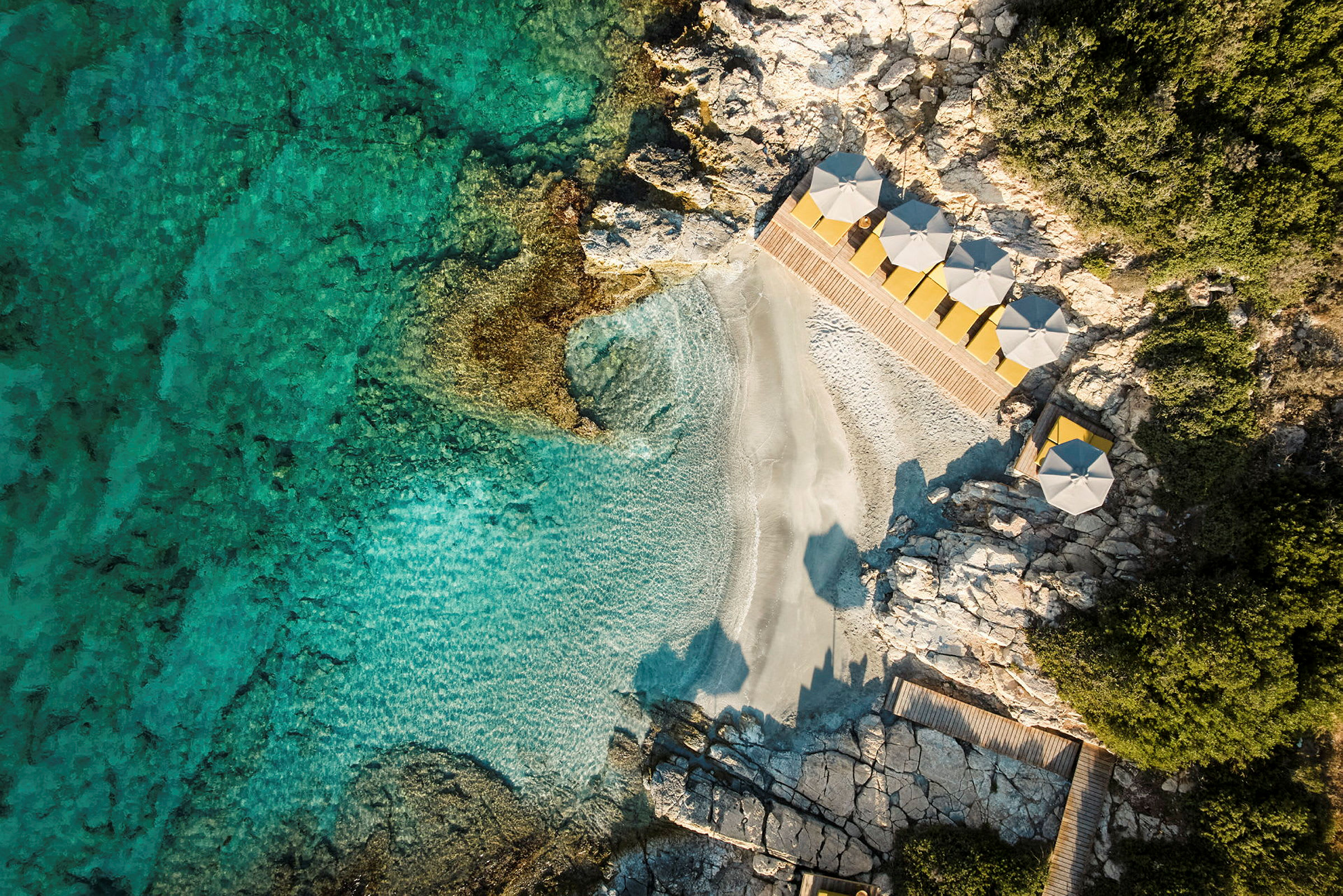 Luftfoto av en liten strand med klart turkist vann, omgitt av klipper og med parasoller ved strandkanten. (AI-generert tekst)