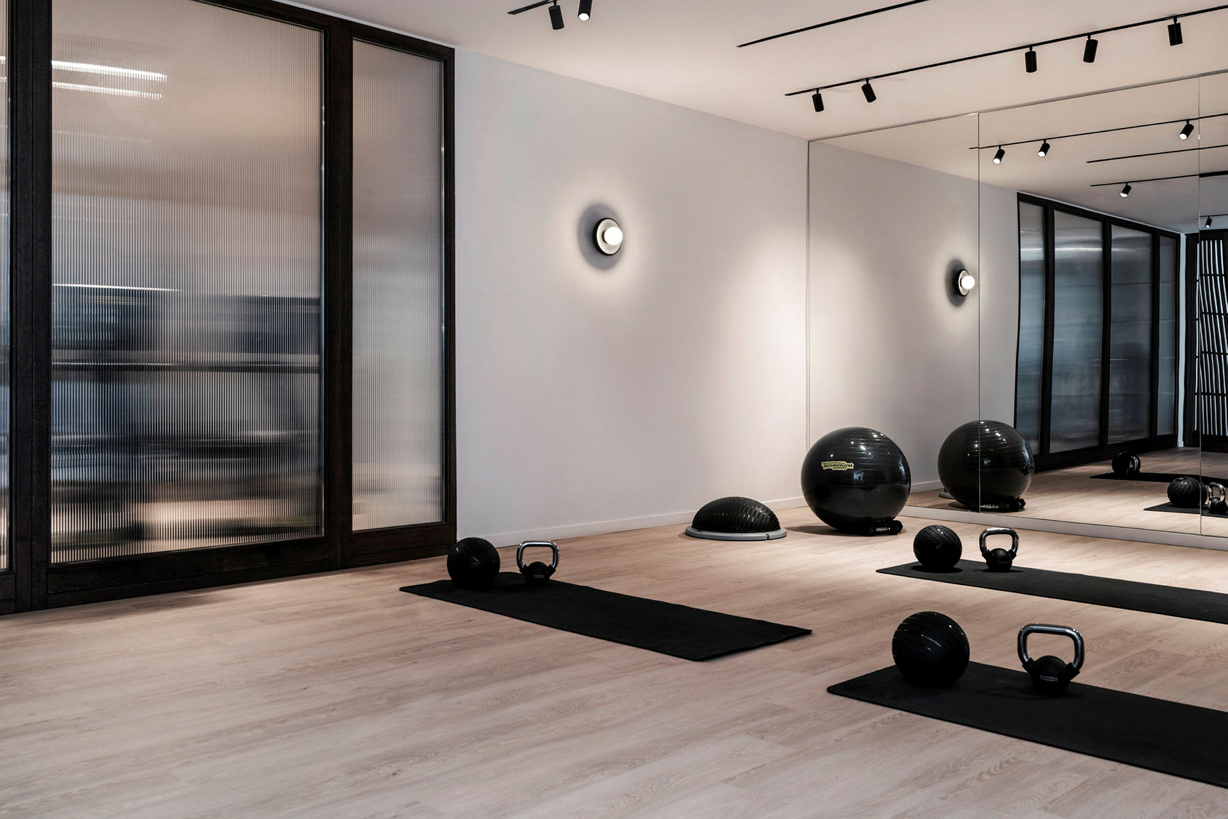 Et moderne treningsstudio med treningsmatter, kettlebells og fitnessballer på et tregulv. Veggen har speil og glasspartier. (AI-generert tekst)