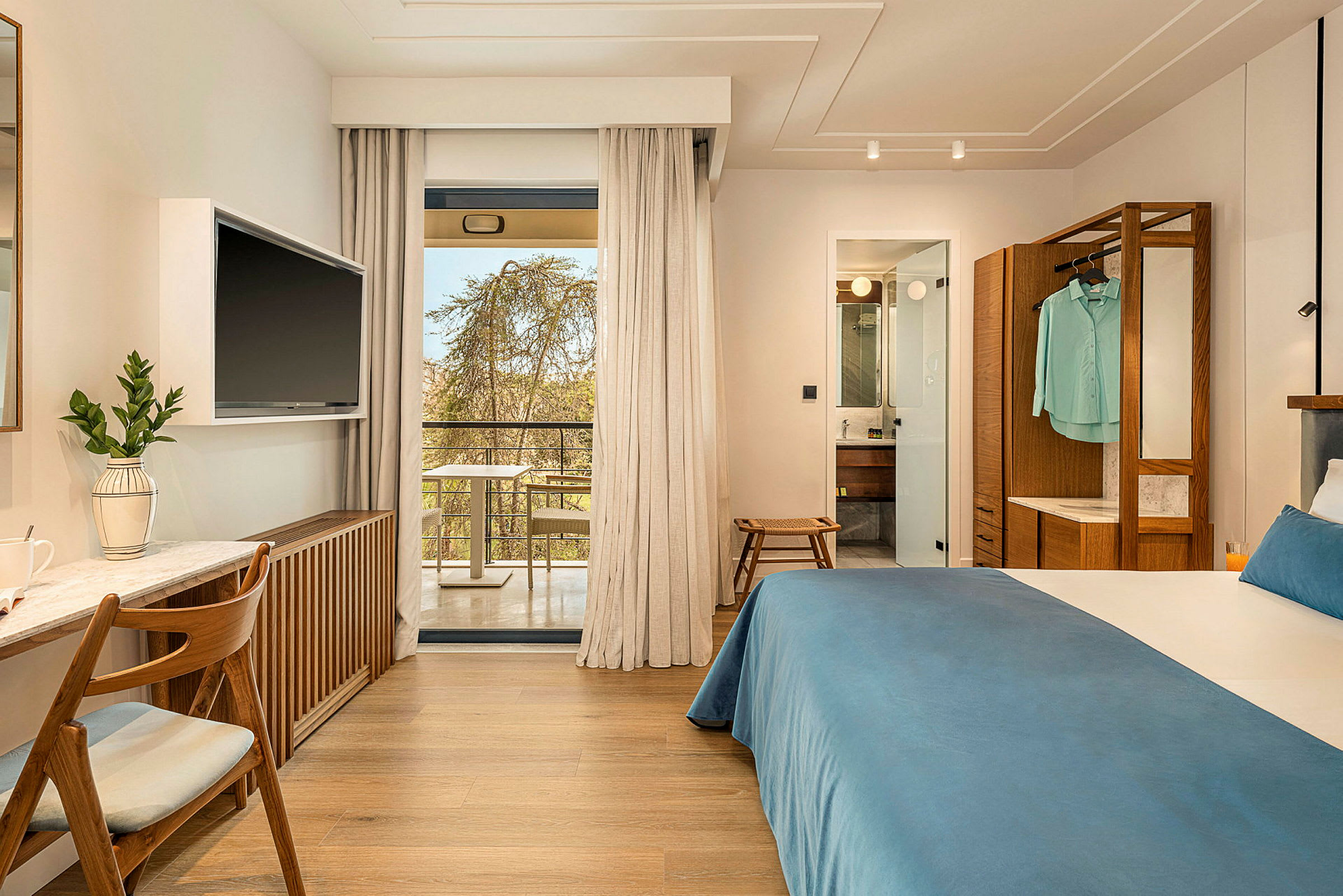Hotellrom med dobbeltseng, skrivebord og TV. Glassdører fører ut til en balkong. Baderommet er synlig gjennom en åpen dør. (AI-generert tekst)