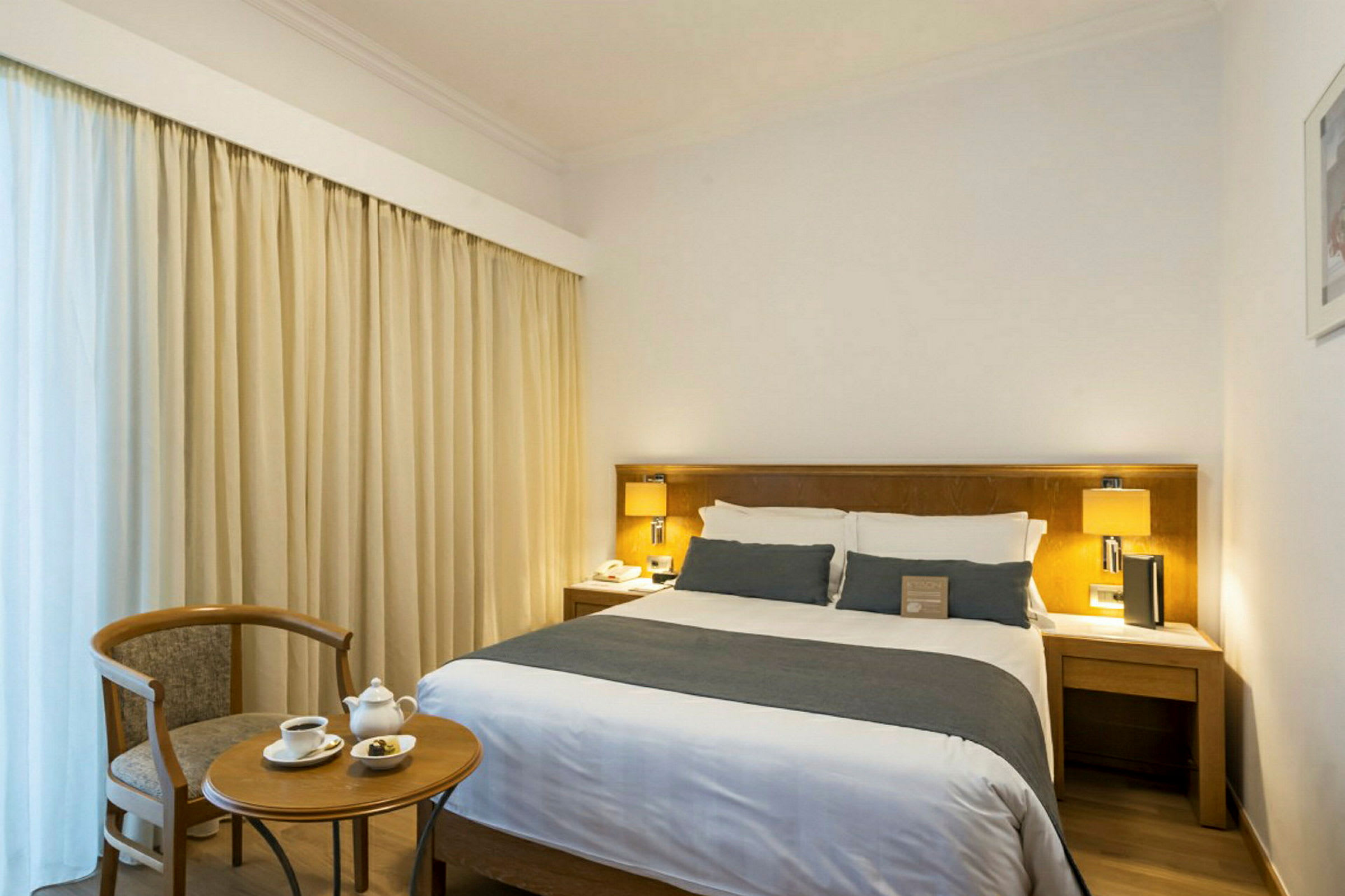 Et hotellrom med en dobbeltseng, nattbord og lampe samt et lite bord med to kopper og en stol. (AI-generert tekst)