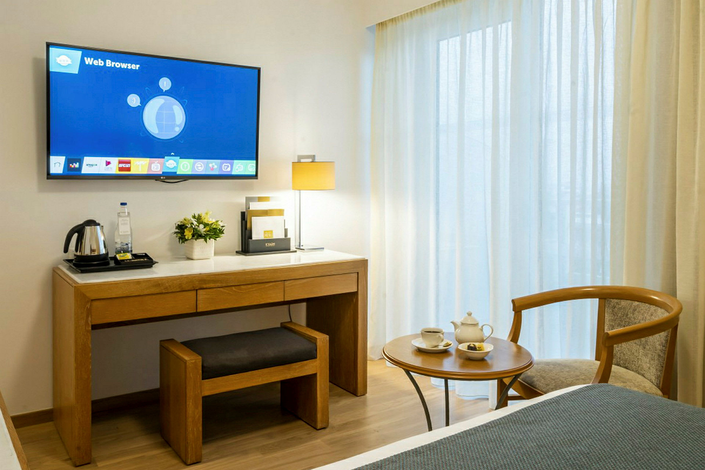 Et hotellrom med en veggmontert TV over et skrivebord med en stol og en vannkoker på toppen. Ved siden av skrivebordet finnes et sidebord med tekopper og en stol. Bakgrunnen inneholder vinduer med lysskjermende gardiner. (AI-generert tekst)