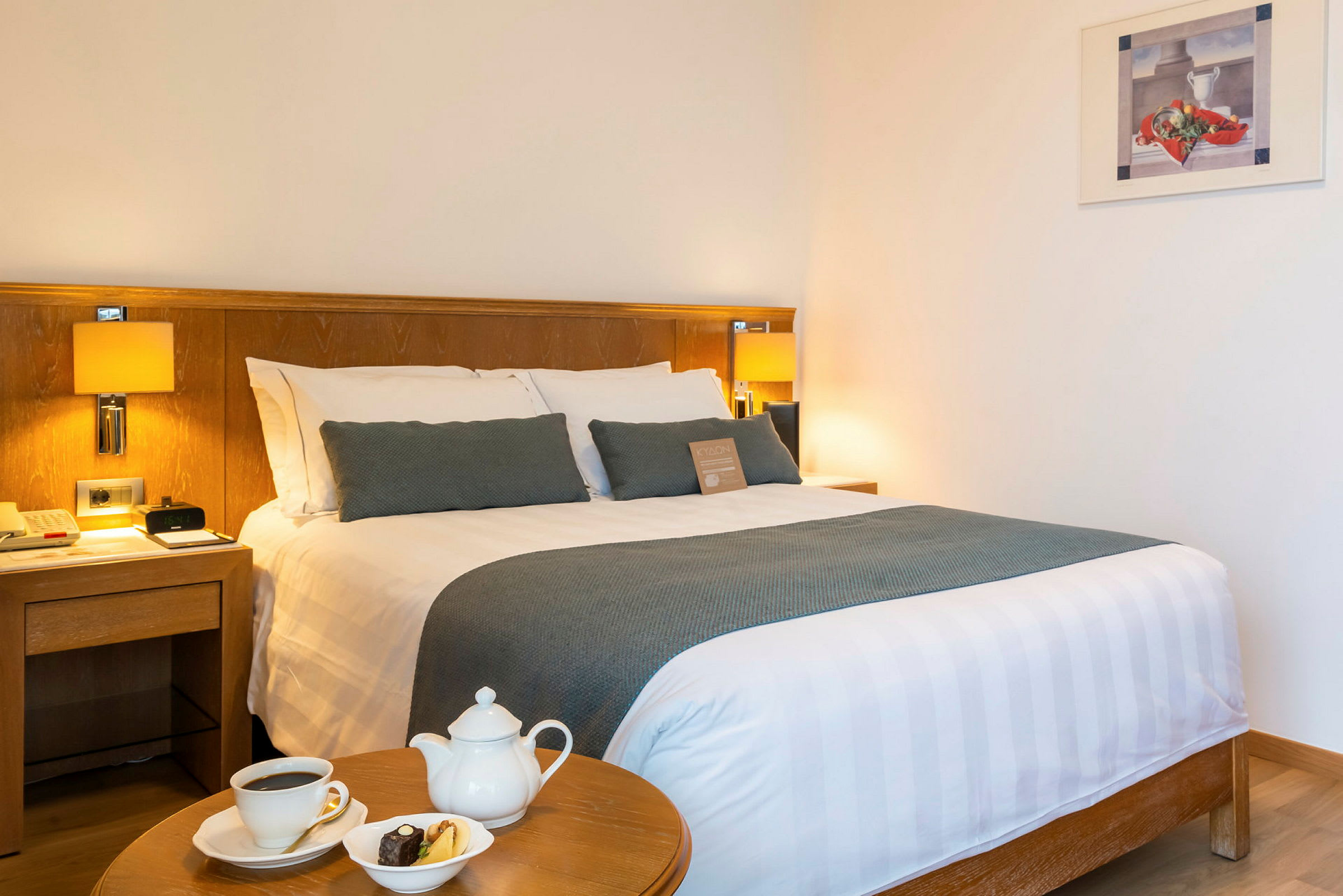 Et hotellrom med en dobbeltseng, nattbord og et bord med en kaffekopp, tekanne og fat med kaker. (AI-generert tekst)