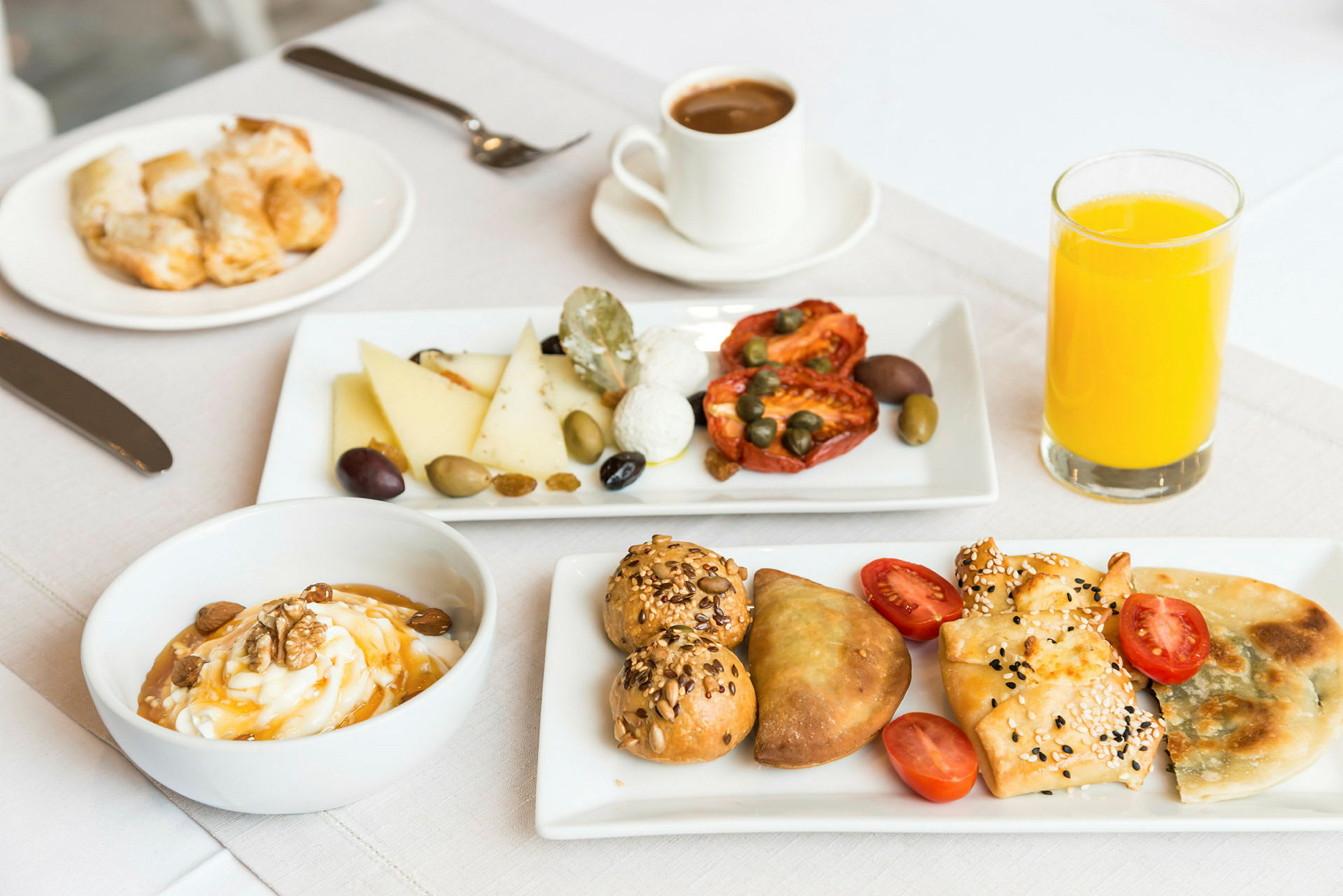 Bildet viser en frokostservering med ulike ingredienser, som brød, ost, soltørkede tomater, oliven og kapers. Det er også en skål med yoghurt, noen bakverk og et glass appelsinjuice. (AI-generert tekst)