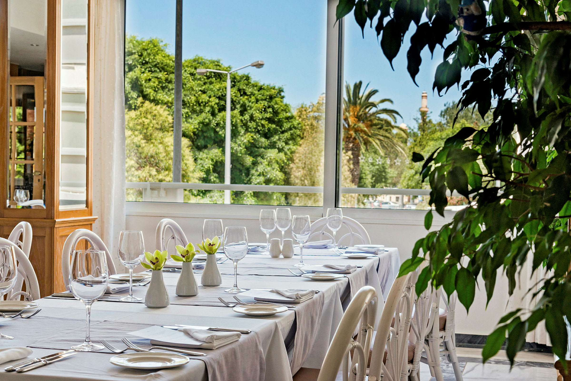 Bildet viser et dekket bord i en restaurant med hvitt porselen og glass. I bakgrunnen er det store vinduer som gir utsikt til frodig natur. (AI-generert tekst)