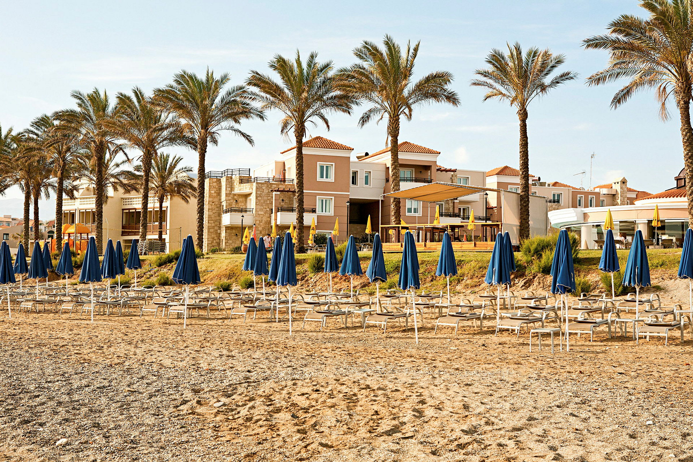 Strand med solsenger og blå parasoller foran hotellbygninger og palmer. (AI-generert tekst)