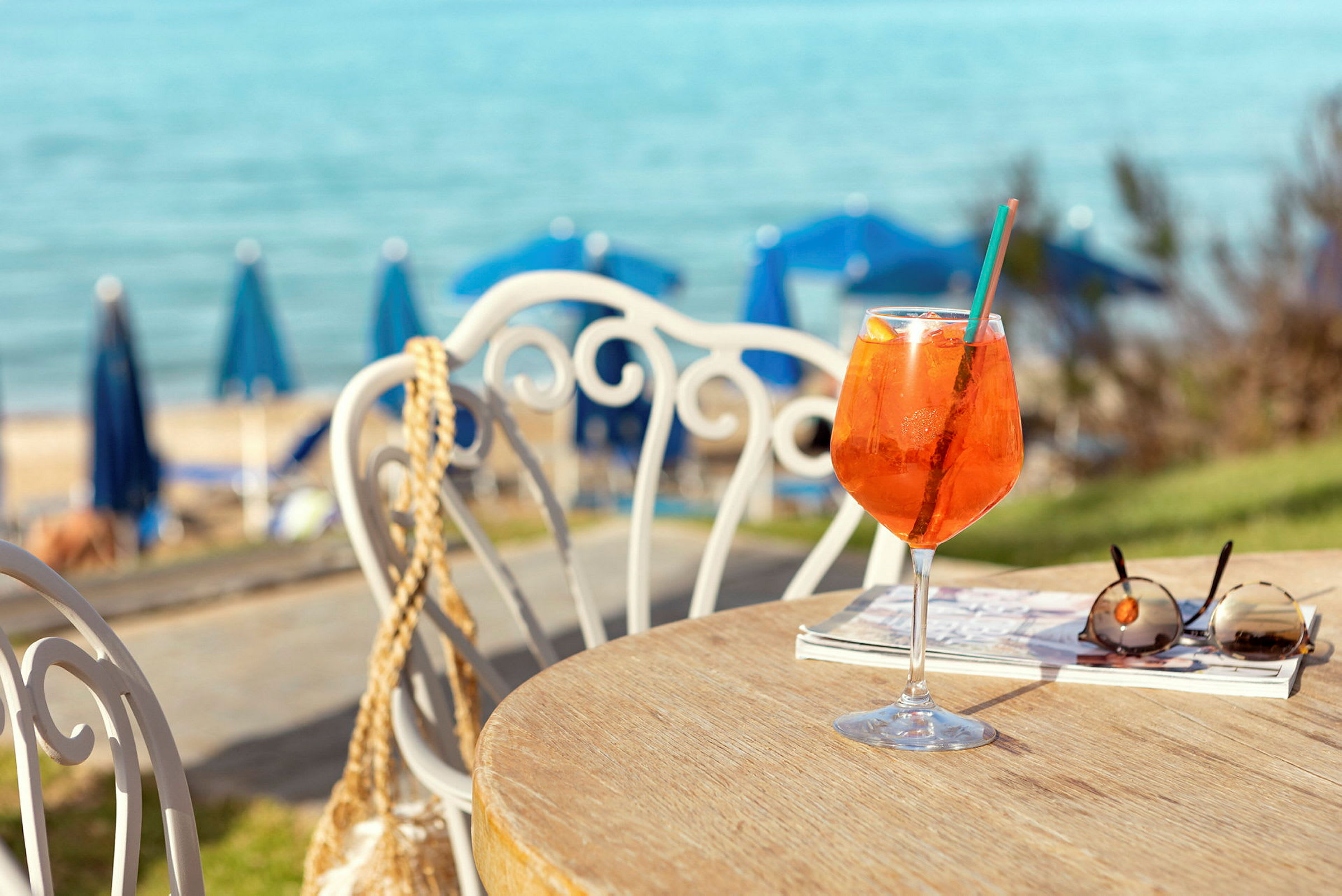 Et bord med et glass appelsindrikk, solbriller og en bok. I bakgrunnen sees en sjø og blå parasoller på stranden. (AI-generert tekst)