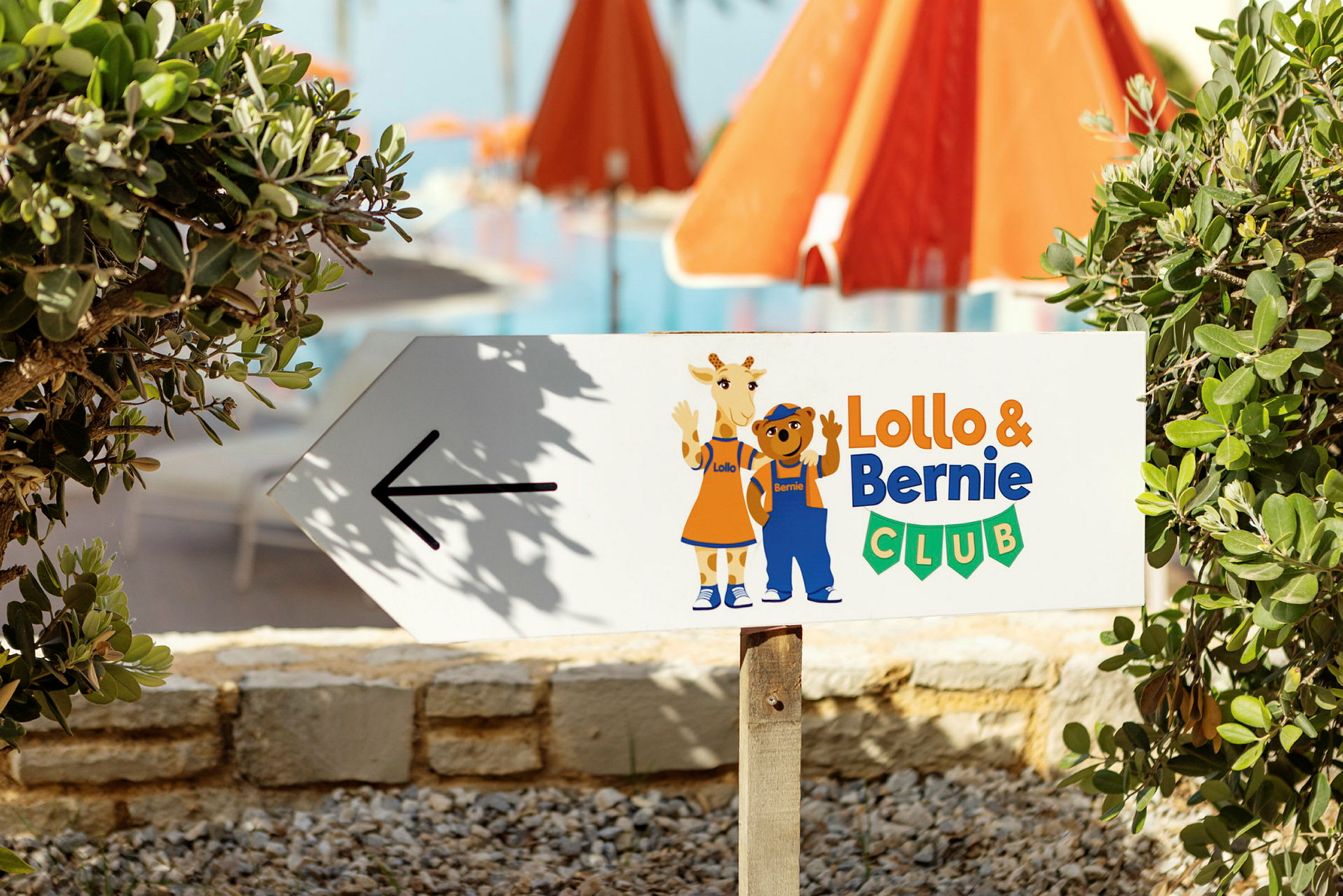 Skilt med pil og teksten 'Lollo & Bernie Club', med illustrasjoner av Lollo og Bernie. Bakgrunn med vegetasjon og røde parasoller. (AI-generert tekst)