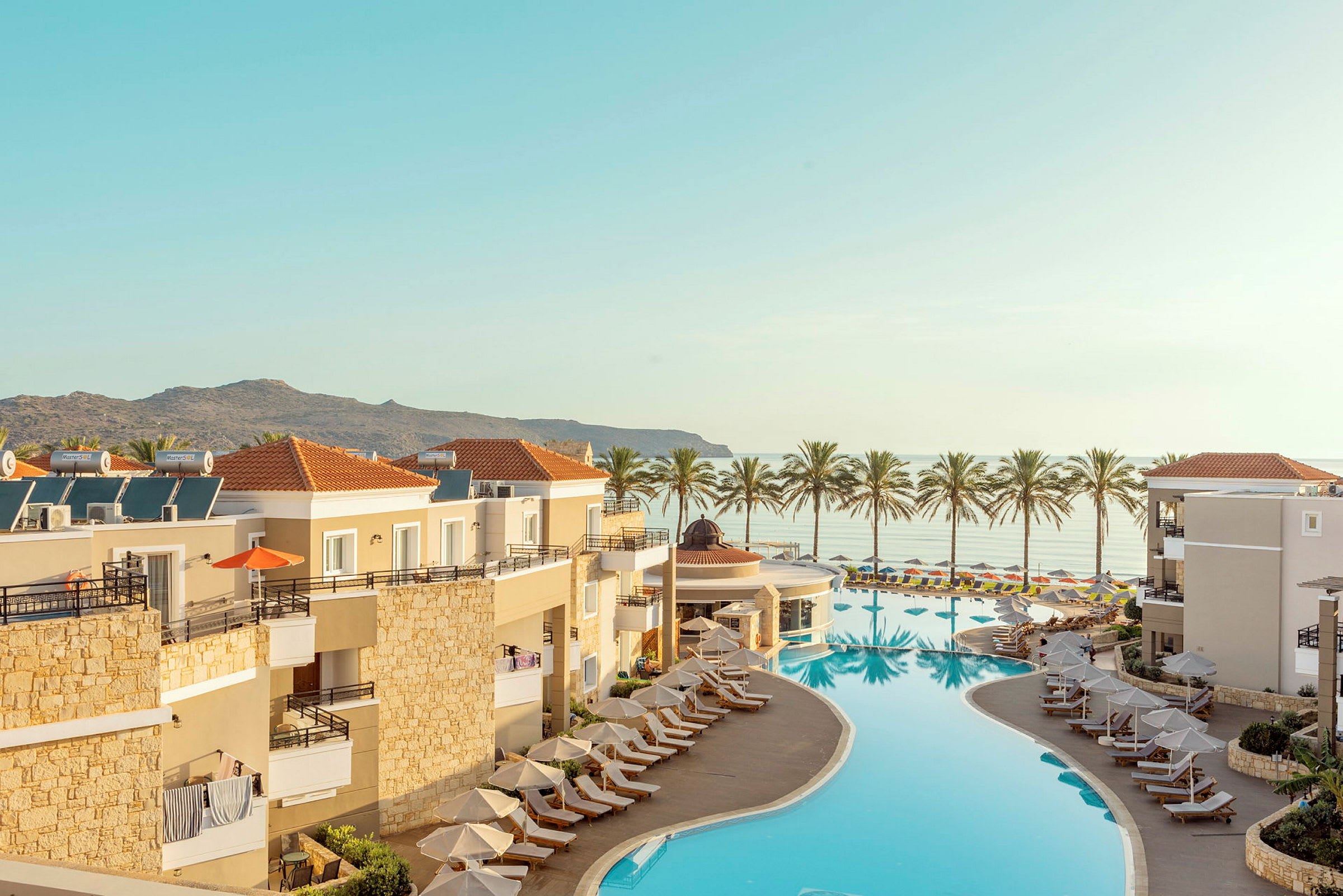 Oversikt over en resort med basseng og solsenger, med havutsikt og palmer i bakgrunnen. (AI-generert tekst)