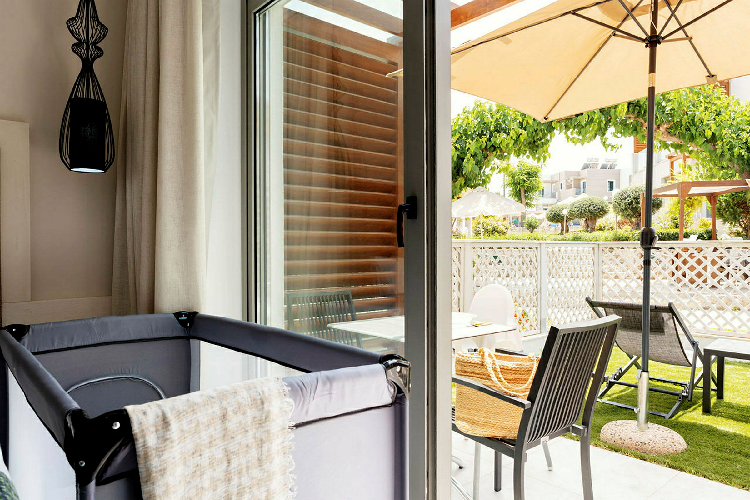 Bilde av et rom med barneseng nær et glassvindu som vender mot en terrasse med utemøbler og parasoll. (AI-generert tekst)