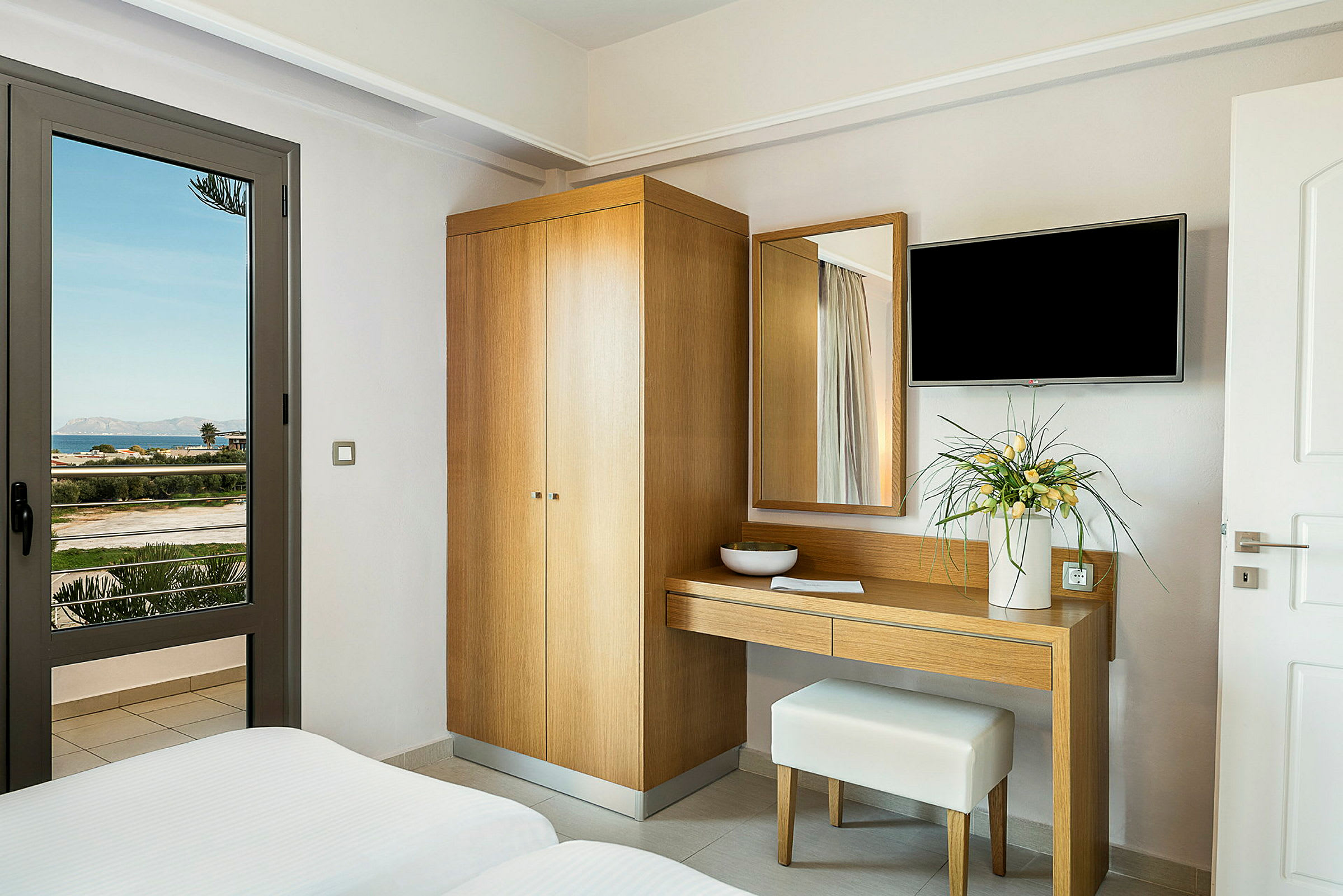 Hotellrom med garderobe, skrivebord og blomsterdekorasjon, samt utsikt over landskap gjennom glassdør. (AI-generert tekst)