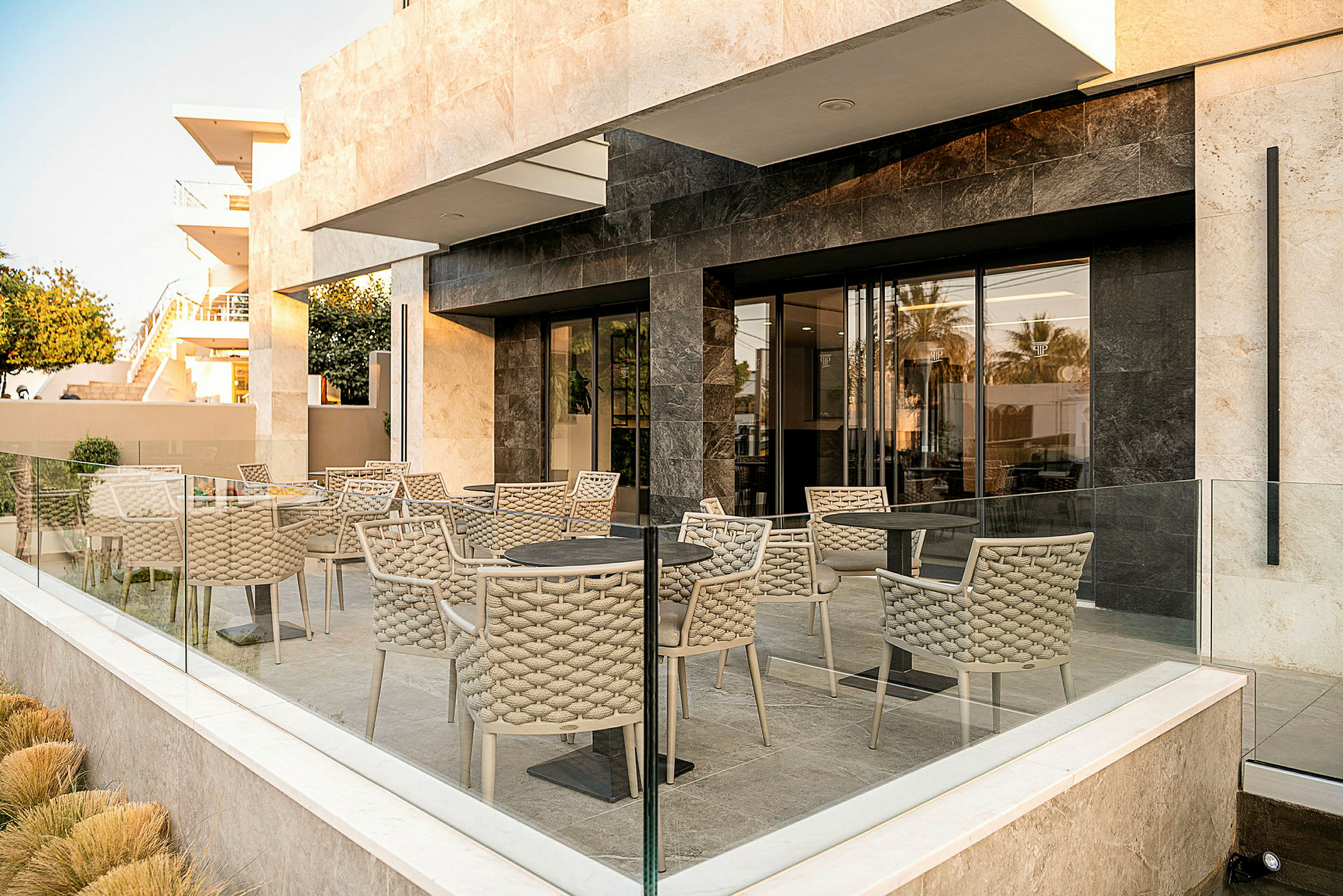 Utendørs terrasse med bord og stoler omgitt av glassrekkverk og moderne bygningseksteriør. (AI-generert tekst)