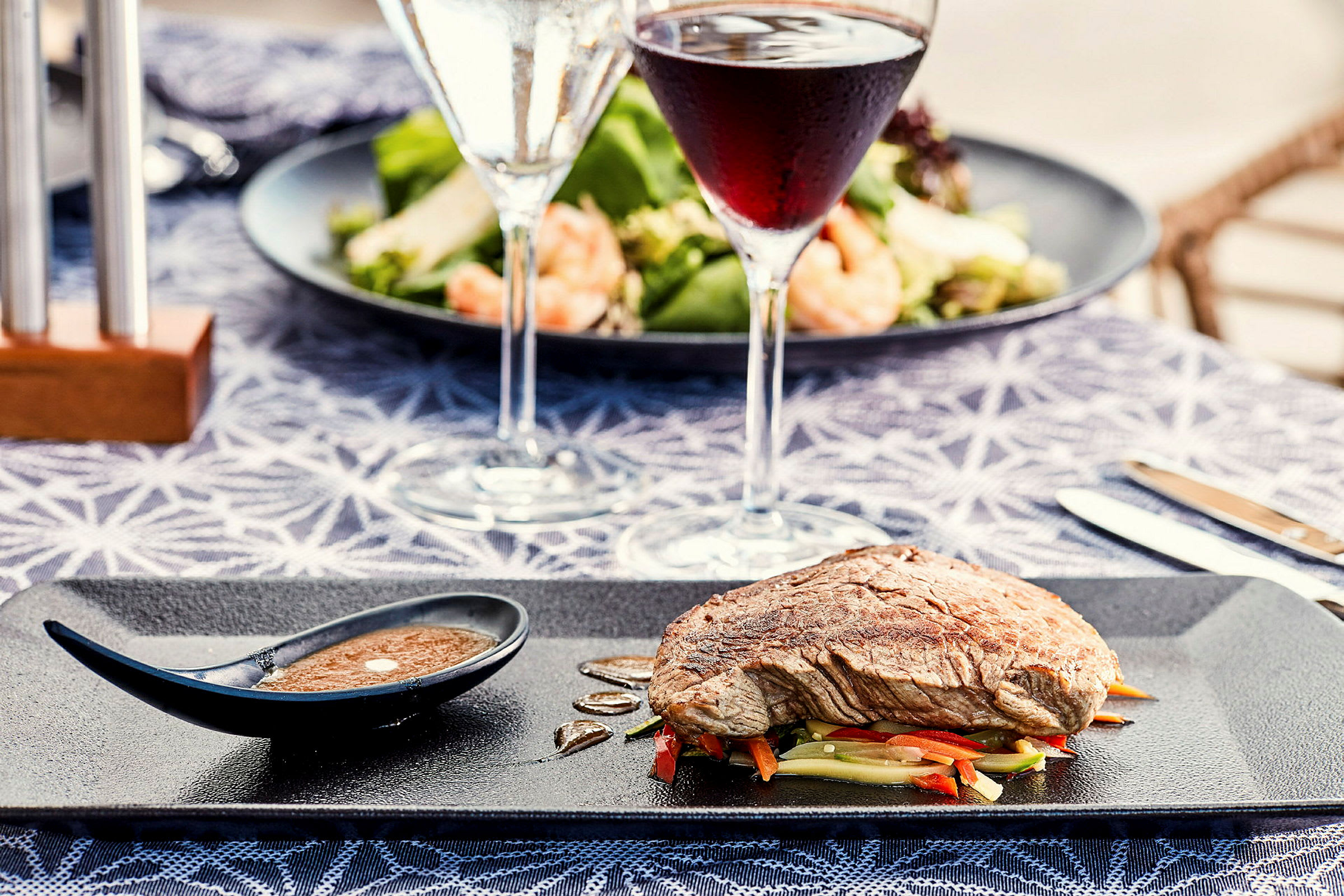En tallerken med kjøtt på grønnsaker og en liten sausbolle, med vinglass og salat i bakgrunnen. (AI-generert tekst)