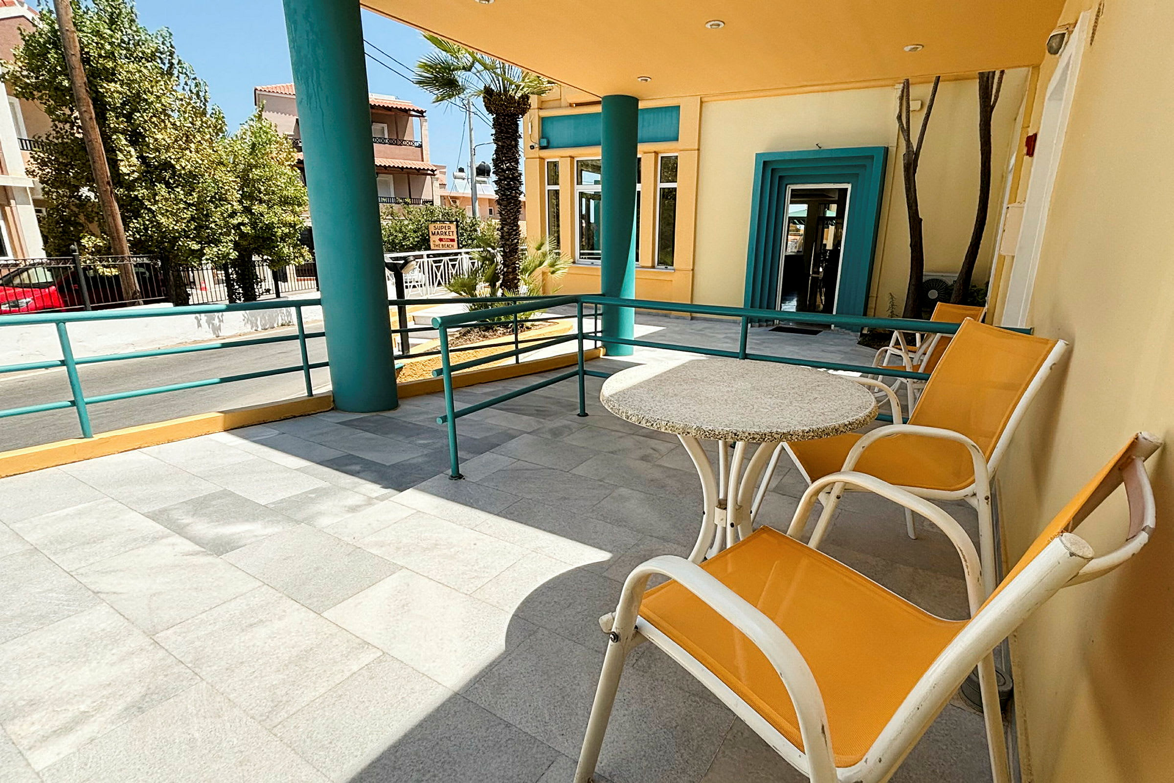 En terrasse med bord og stoler, omgitt av fargerike bygninger og trær. En vei er synlig i bakgrunnen. (AI-generert tekst)