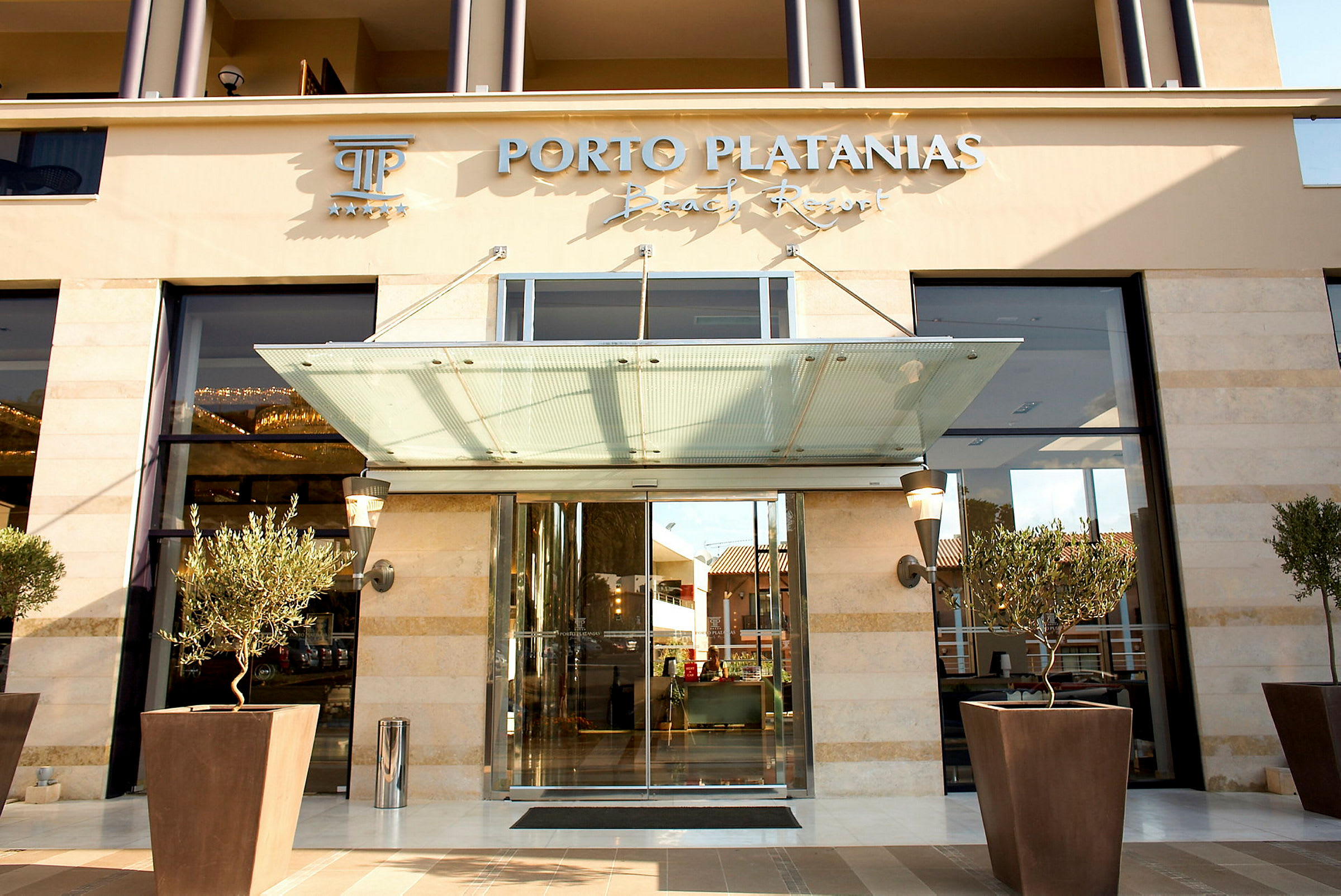 Inngangen til Porto Platanias Beach Resort, med glassdører og beplantede oliventrær foran. (AI-generert tekst)