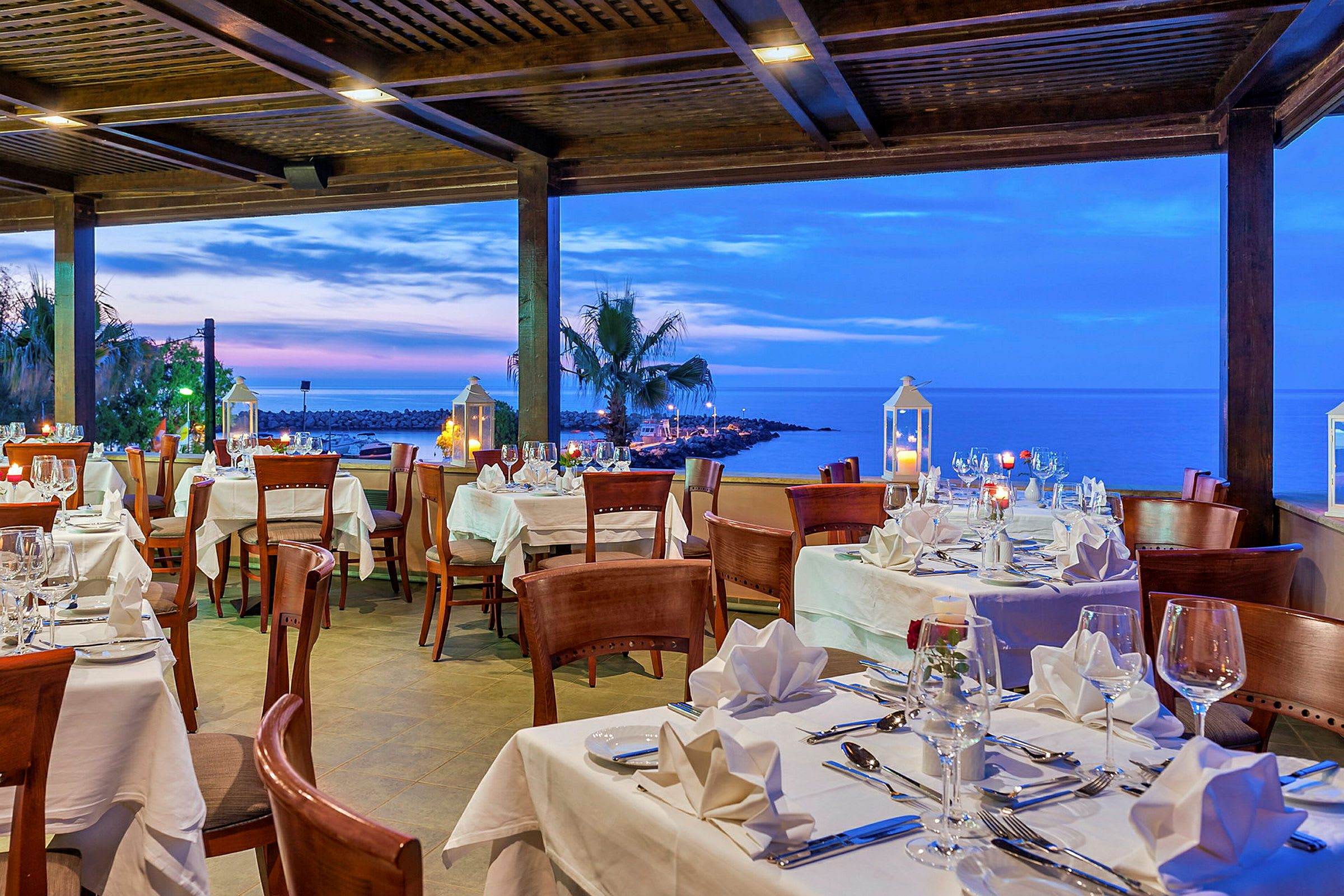 En restaurant med dekkede bord med hvite duker, med utsikt over havet og solnedgangen. (AI-generert tekst)