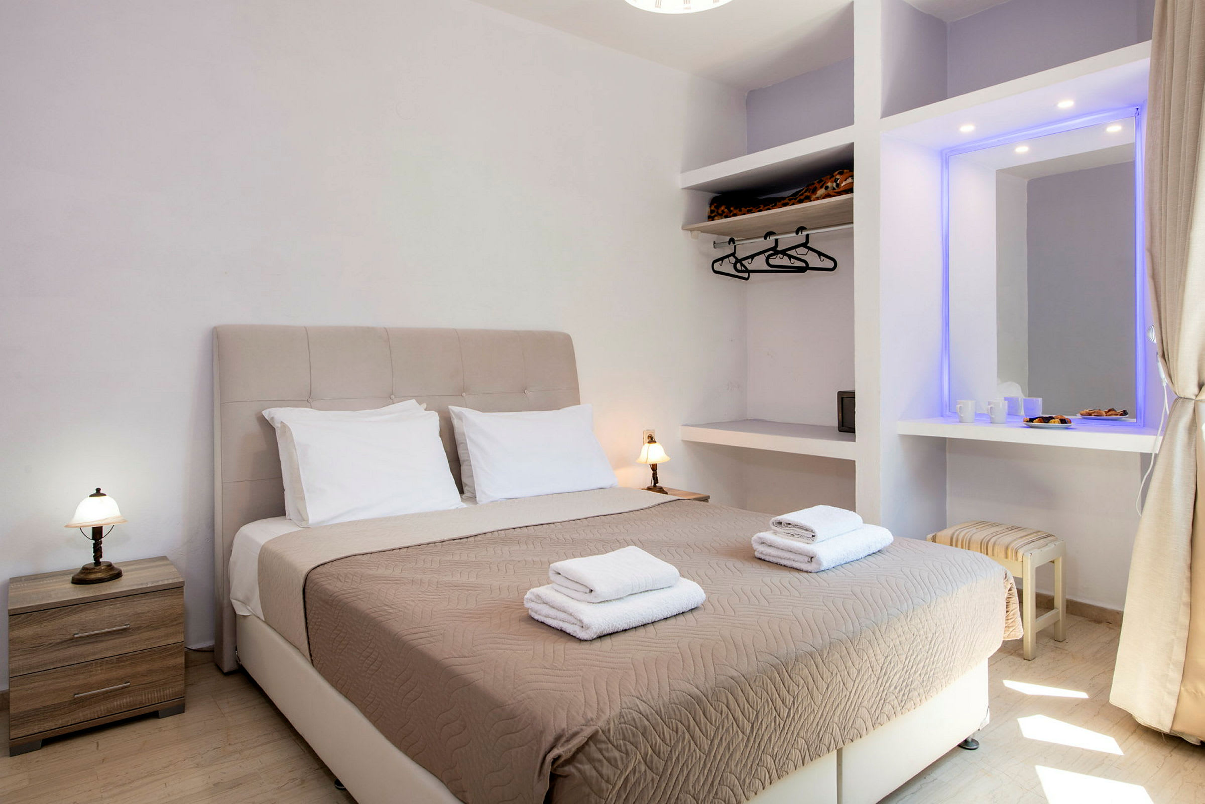 Et hotellrom med en dobbeltseng utstyrt med beige sengeteppe og to sett med håndklær, samt sidebord og garderobe. (AI-generert tekst)