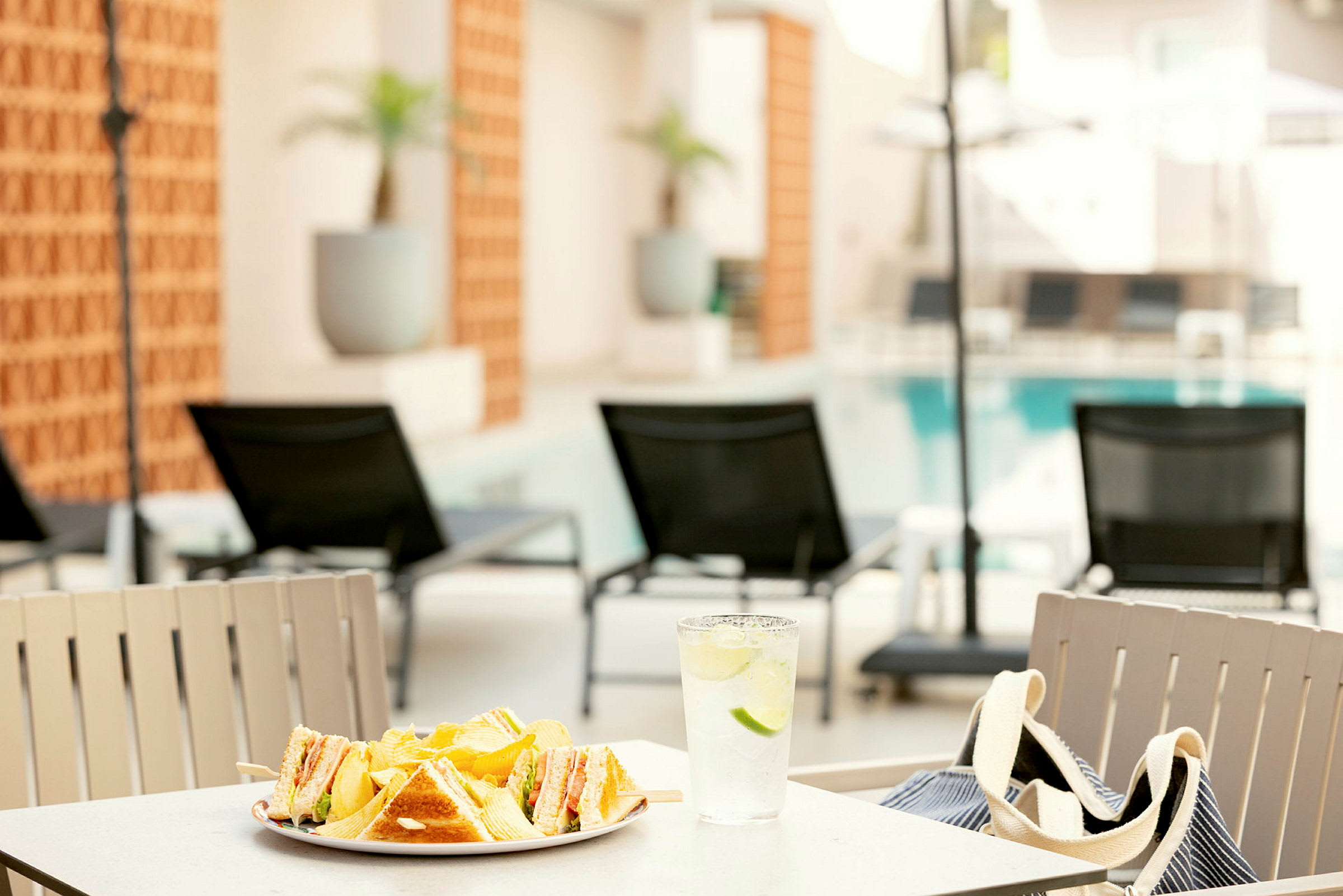En tallerken med smørbrød og chips samt et glass vann med lime står på et bord ved et basseng med solsenger i bakgrunnen. (AI-generert tekst)