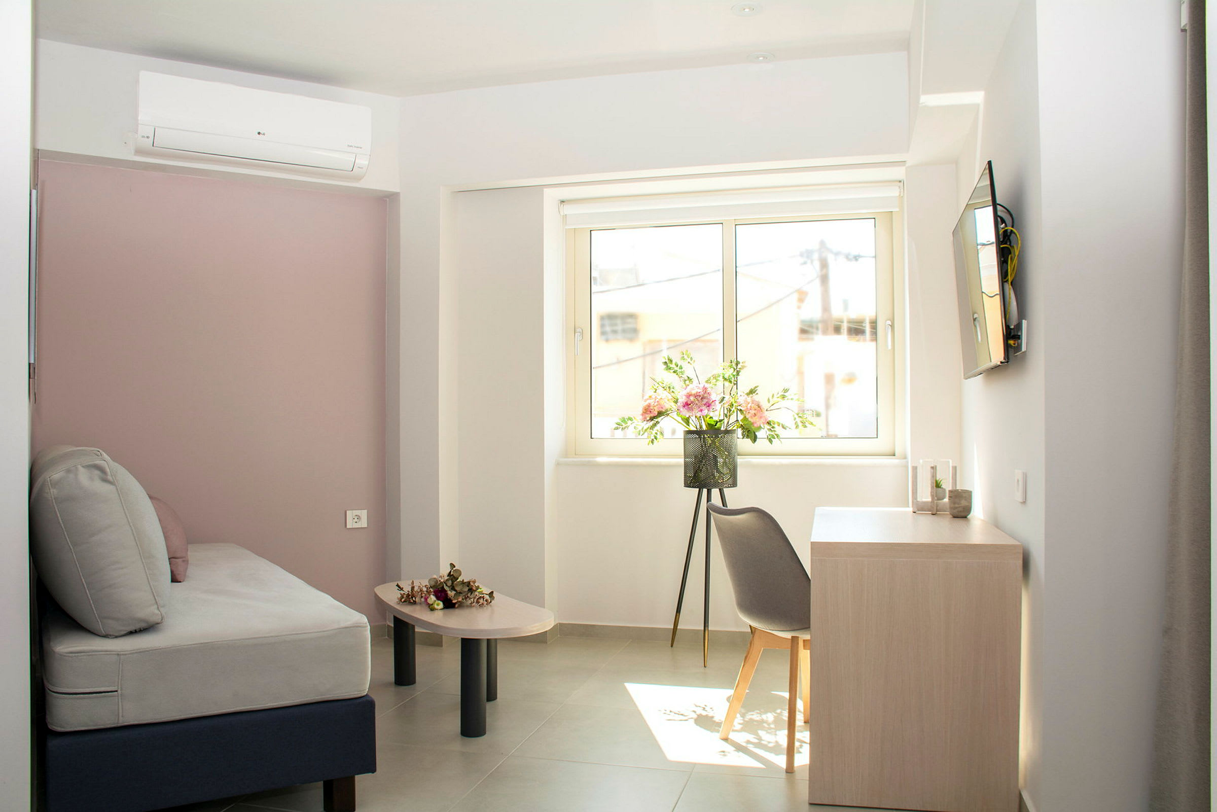 Et lyst hotellrom med grå sofa, blomster på bordet og vindusutsikt. (AI-generert tekst)