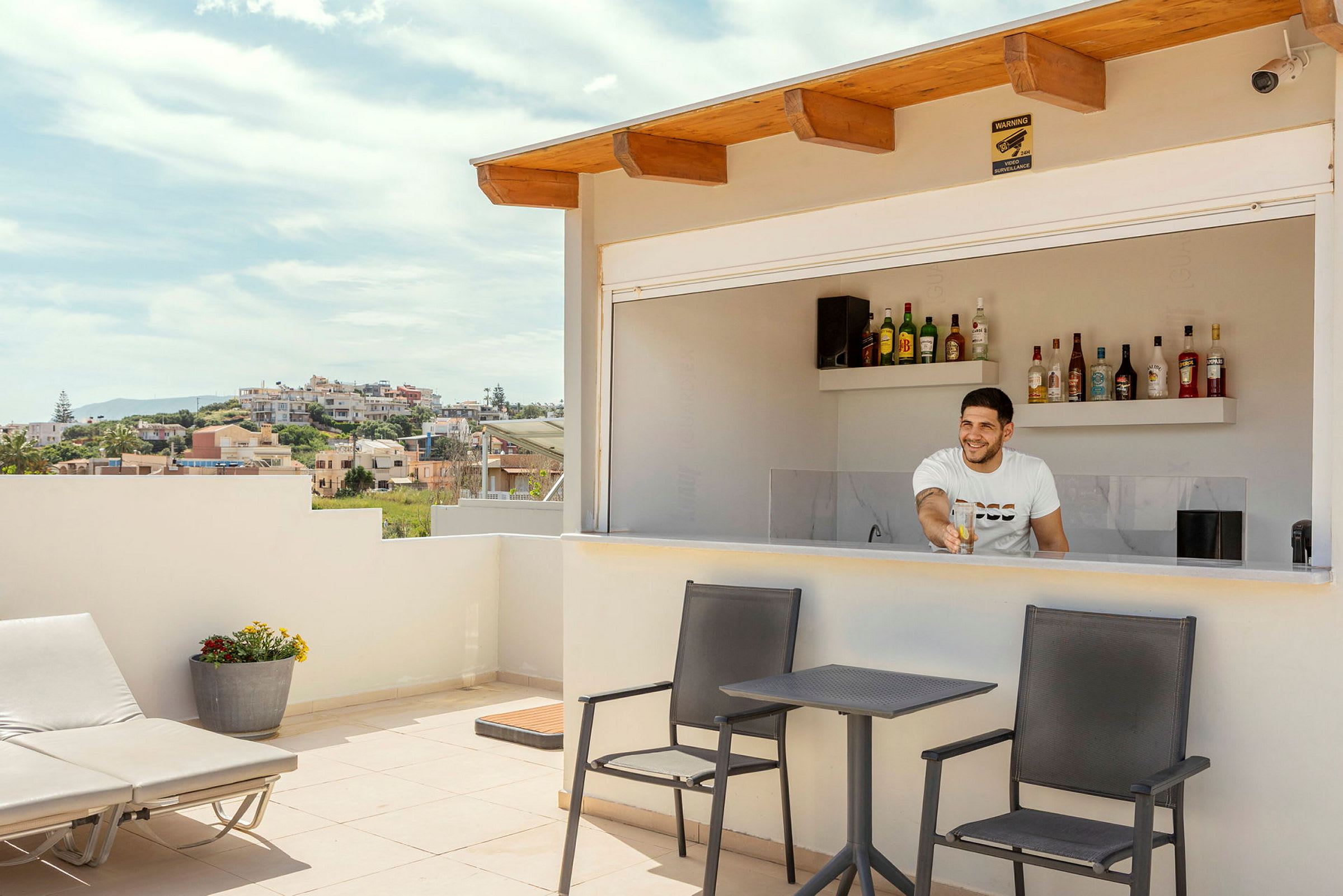 En terrasse med en bar der en person står bak disken. Foran baren er det bord med stoler og en solseng. (AI-generert tekst)