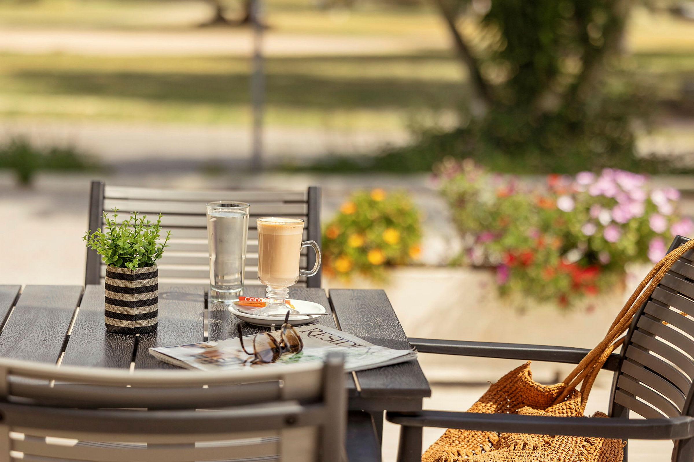 En utendørs servering med et bord som har en kaffekopp, et glass vann, en avis og solbriller. Bakgrunnen viser en frodig miljø og blomster. (AI-generert tekst)