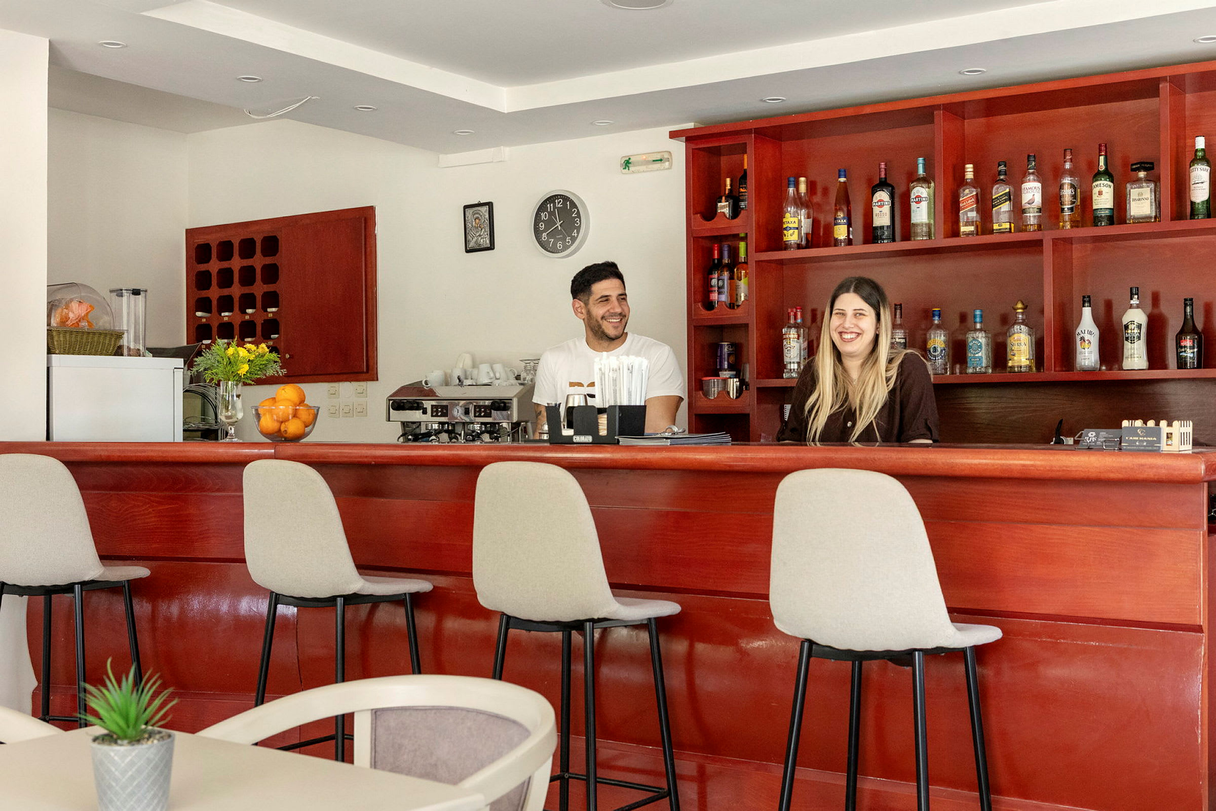 En bar med trepanel og stoler, flasker er plassert på hyllene, og to personer står bak baren. (AI-generert tekst)