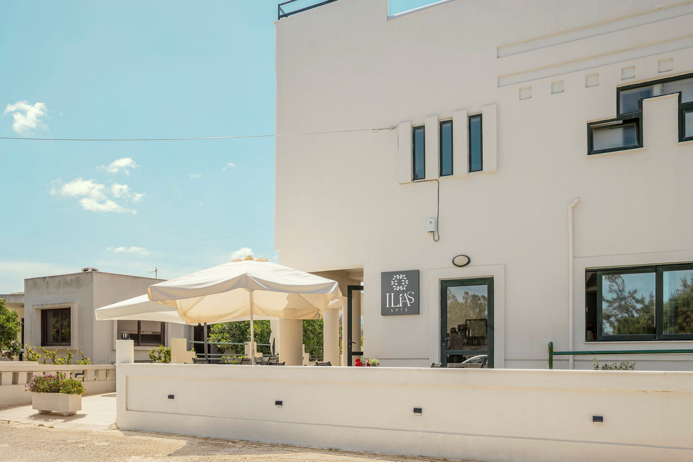 Bilde av en hvit bygning med flere vinduer og et skilt som sier 'Ilias Apts'. Foran bygningen er en uteplass med en stor parasoll og blomsterkasser. (AI-generert tekst)