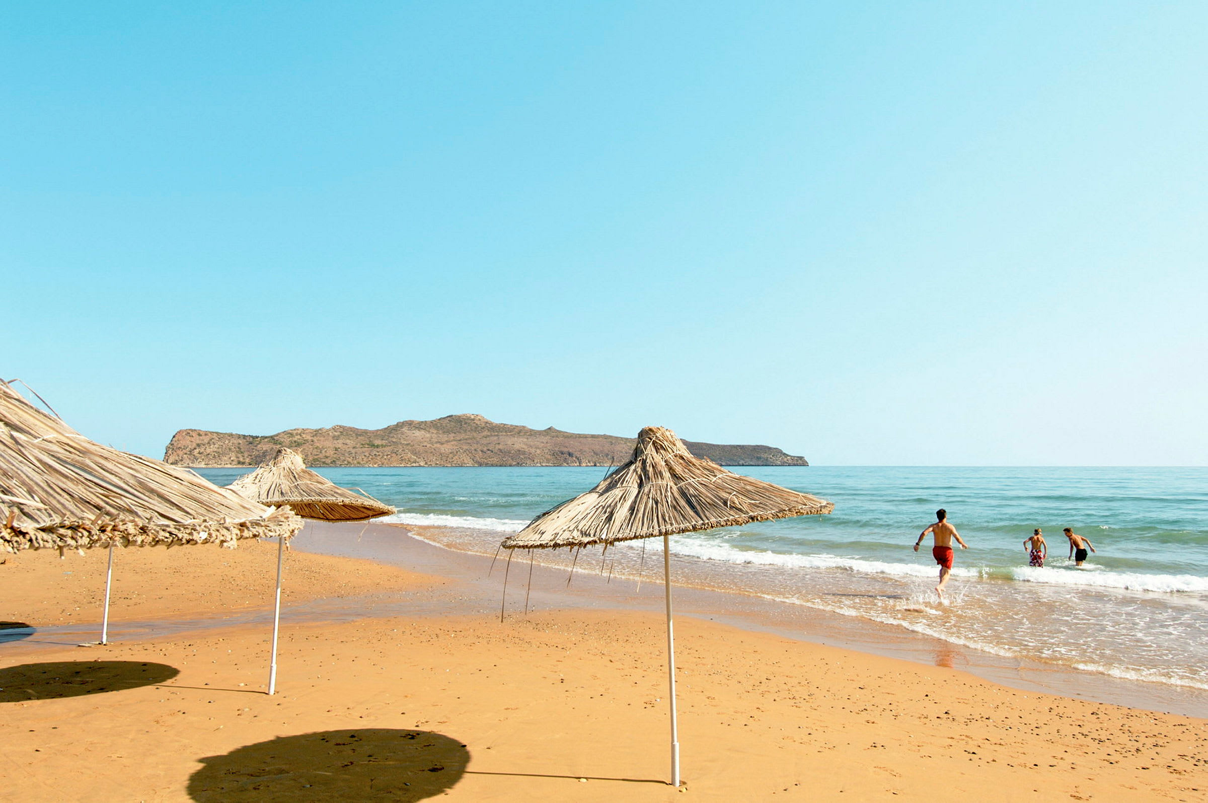 Strand med parasoller av siv og mennesker som bader i havet. (AI-generert tekst)