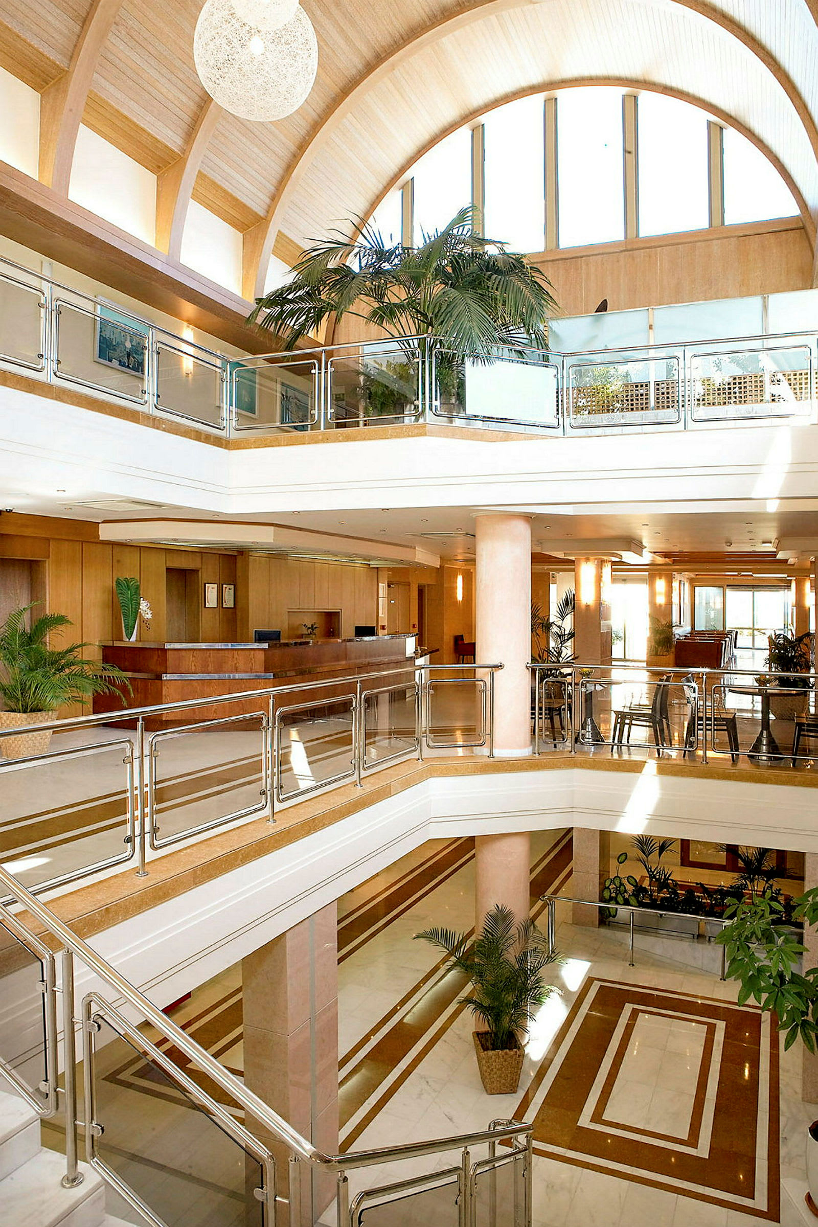 Interiøret i en moderne hotellobby med flere etasjer. Glassrekkverk, tretenger og store palmer er synlige sammen med en resepsjon i bakgrunnen. (AI-generert tekst)