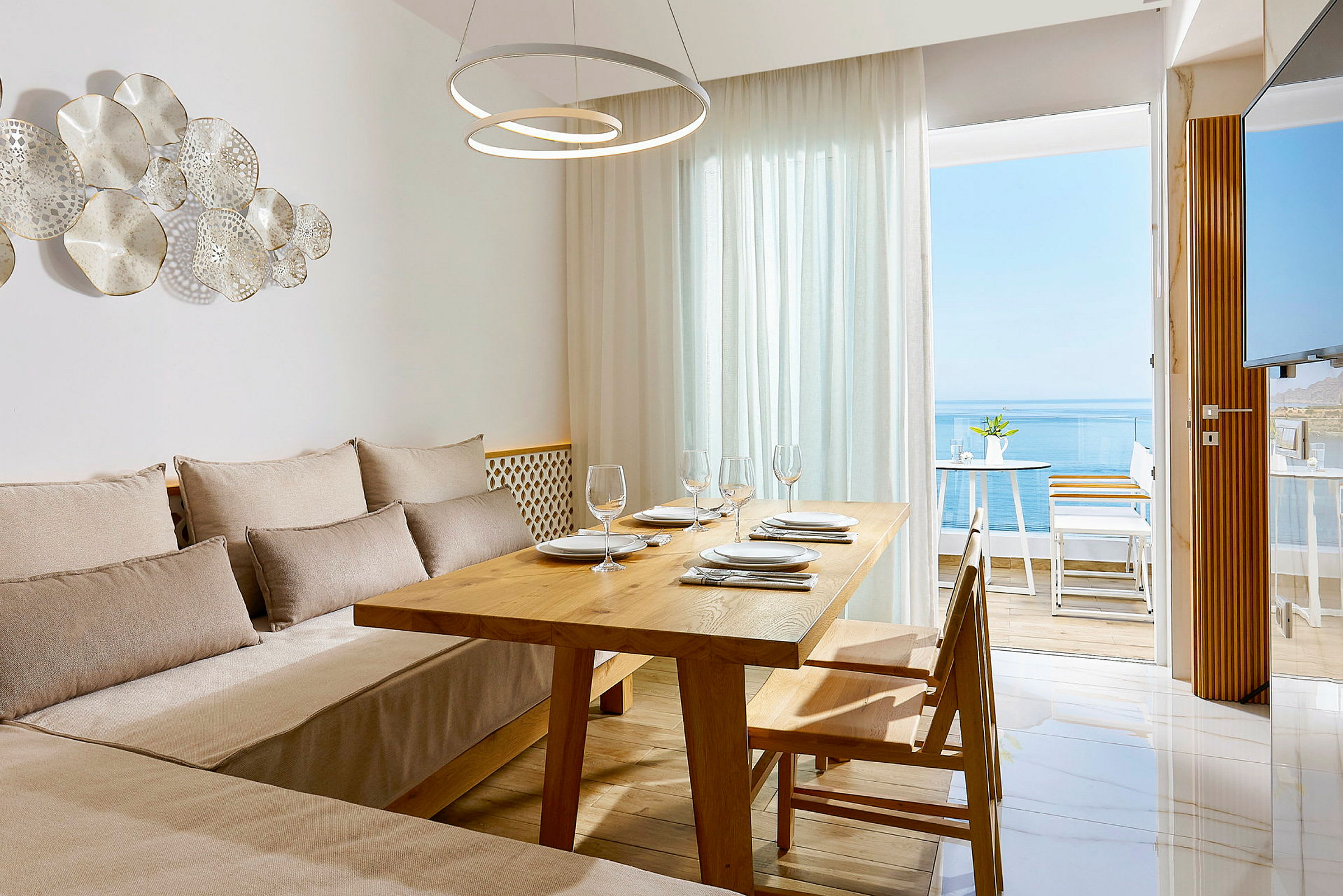 Et lyst og moderne spisestue med et bord av tre, stoler, beige sofaer og en glassdør som åpner opp til en møblert balkong med havutsikt. (AI-generert tekst)
