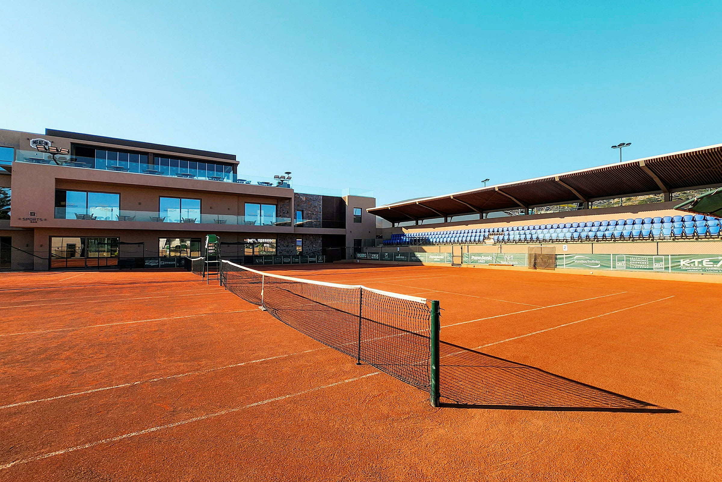 En tennisbane med rød grus og nett foran en bygning og tribuner. (AI-generert tekst)