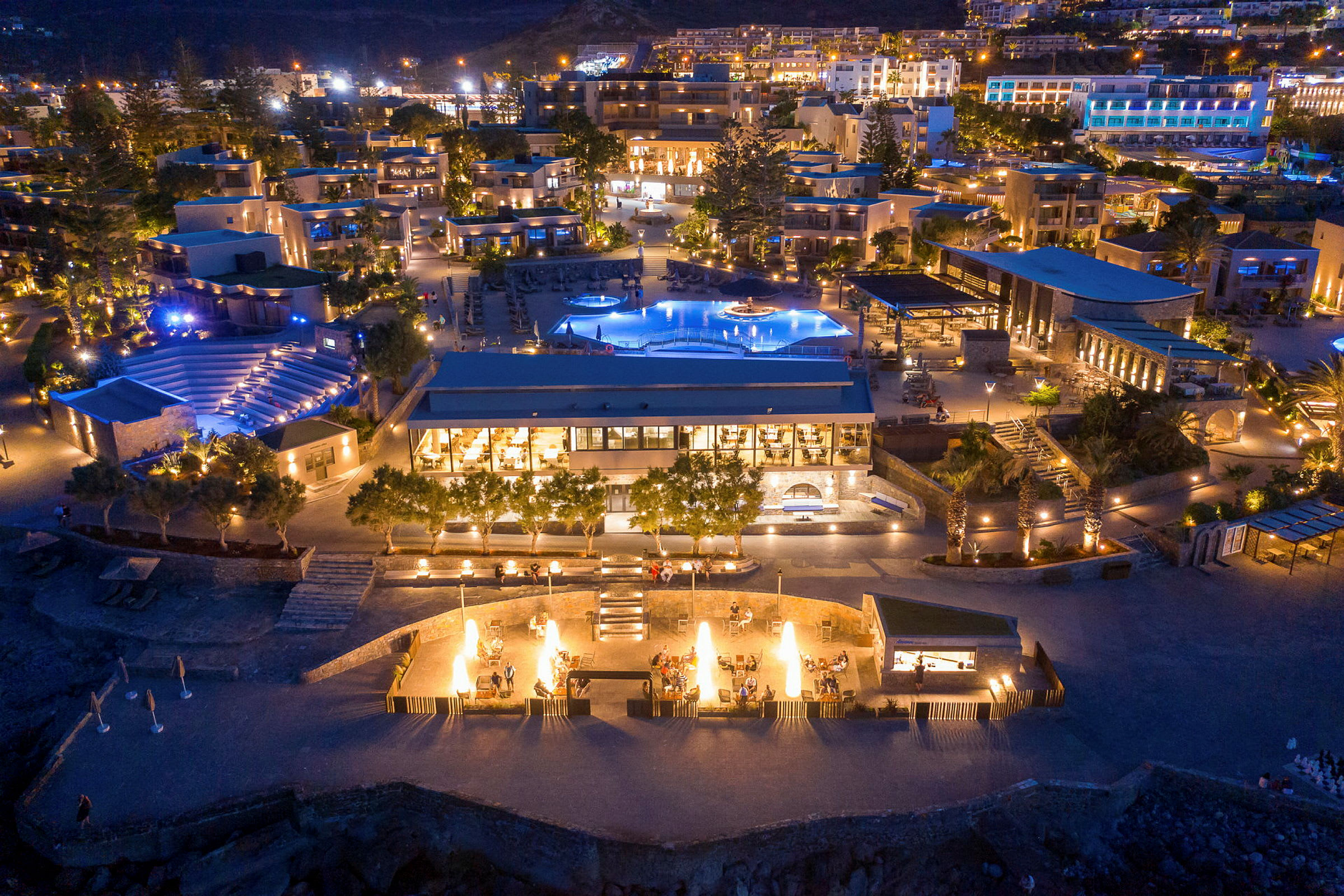 En resort med flere bygninger, bassenger og utendørsområder opplyst om kvelden. (AI-generert tekst)