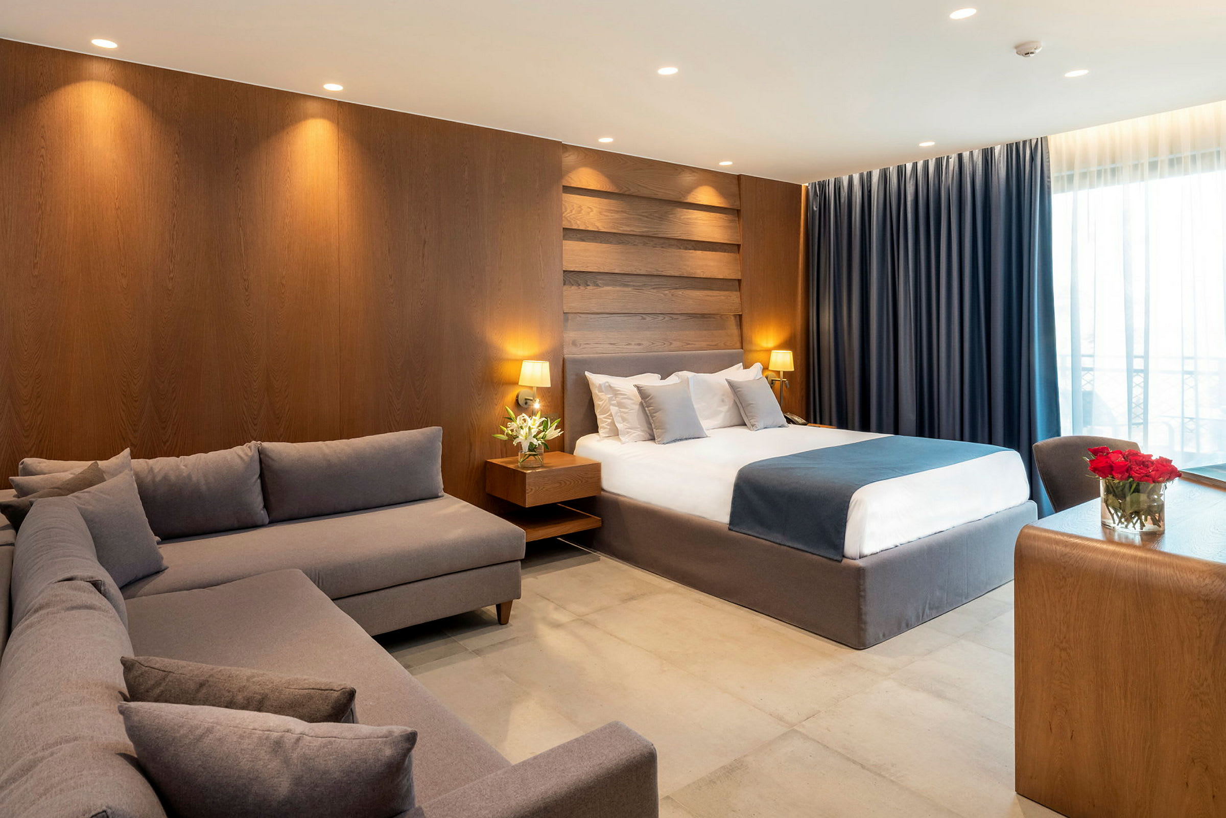 Et hotellrom med en stor seng, en grå sofa og et skrivebord av tre. (AI-generert tekst)