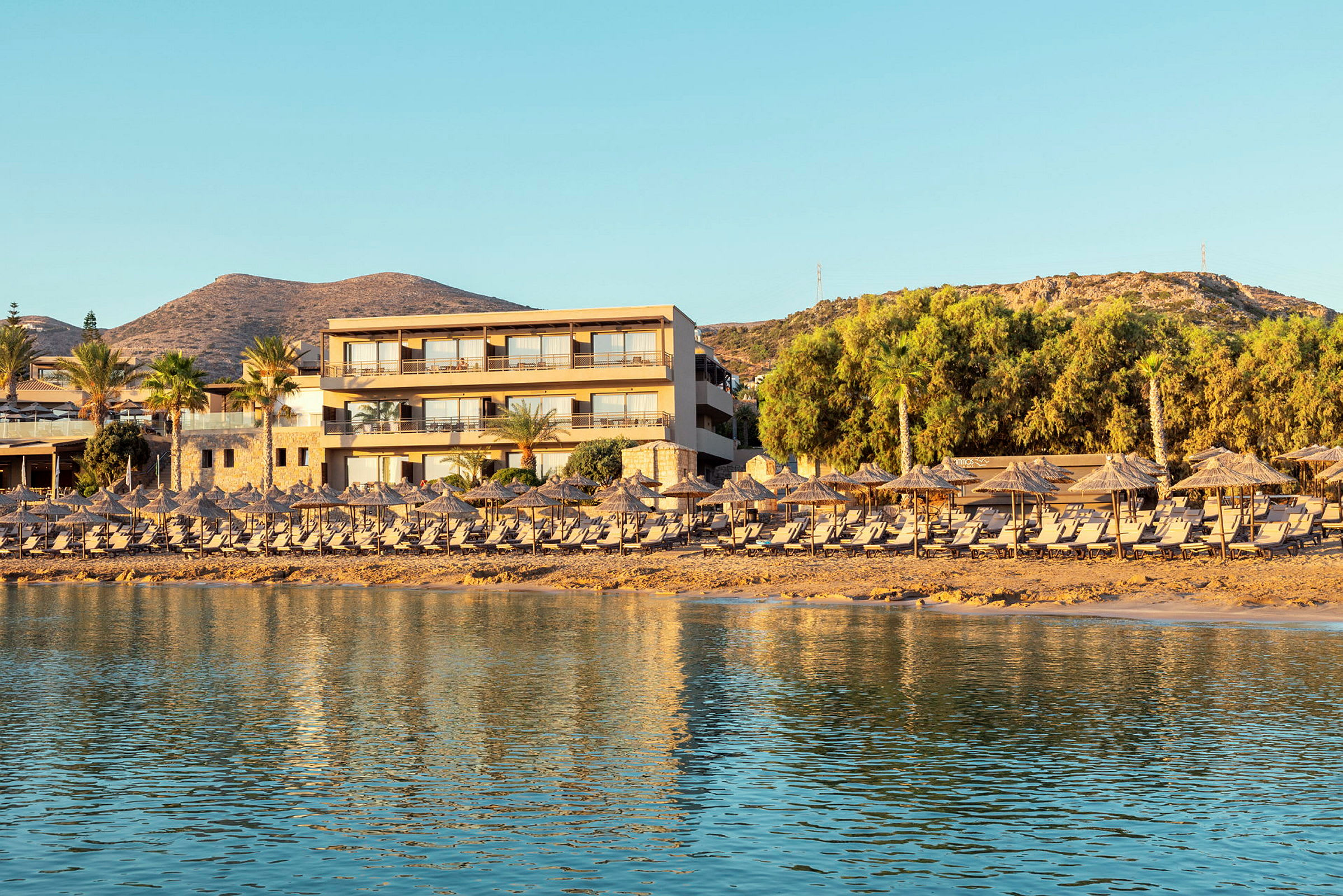En ferieanlegg ved stranden med solsenger og parasoller i forgrunnen og et hotell bak. (AI-generert tekst)